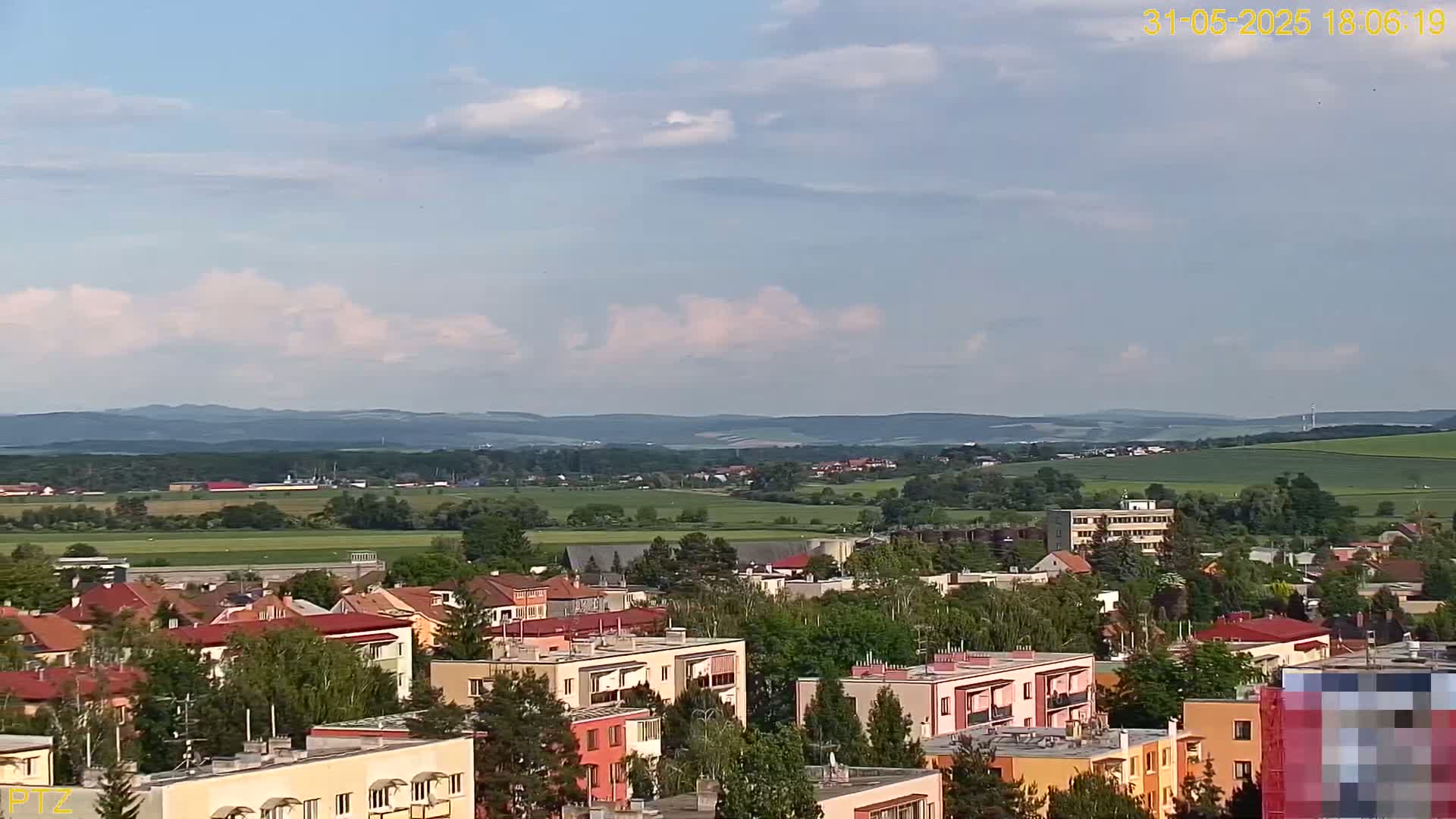 A partly cloudy day reveals a view of a town nestled amongst green fields and distant rolling hills.