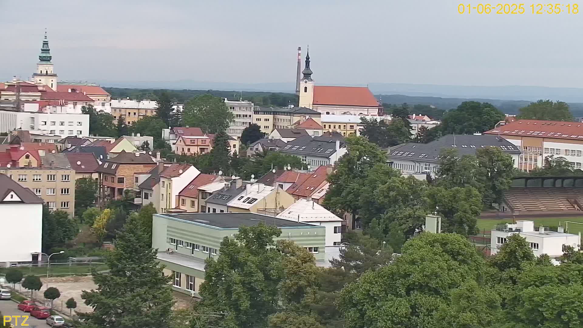 An elevated view of a town on an overcast day, showing a mix of residential buildings, a church steeple, and a stadium nestled among green trees.