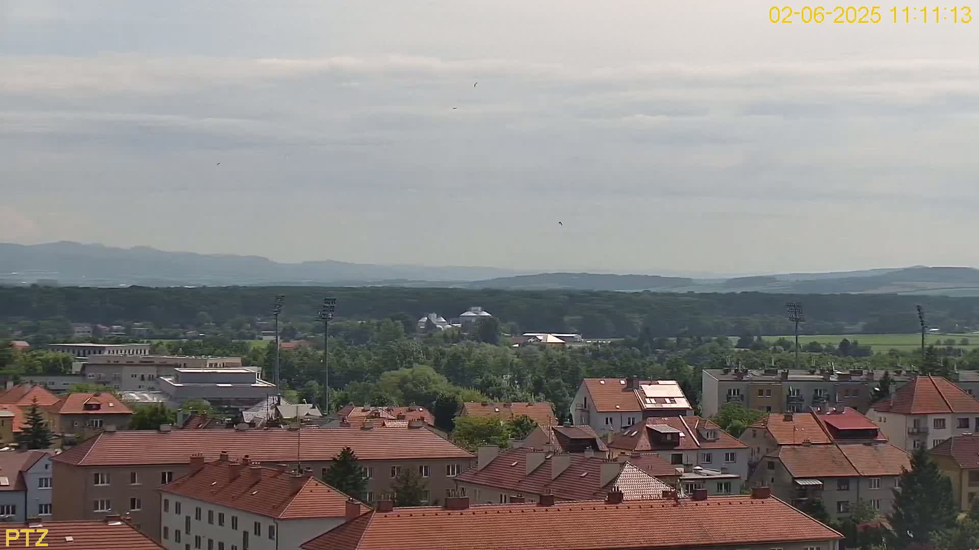 A mostly cloudy day reveals a sprawling town of red-roofed buildings nestled amongst green trees, with distant mountains visible on the horizon.