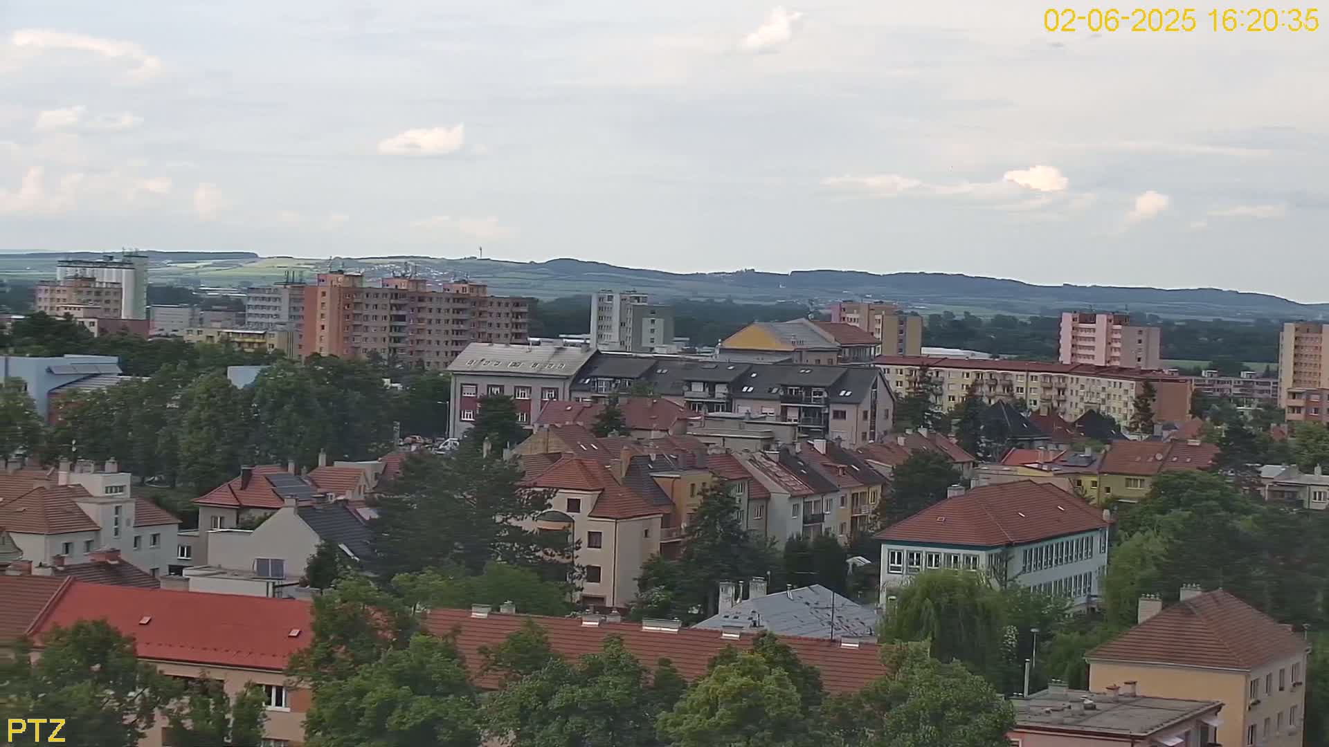 A mostly cloudy day reveals a cityscape of multi-story buildings with red-tiled roofs, interspersed with green trees and a distant hilly landscape.