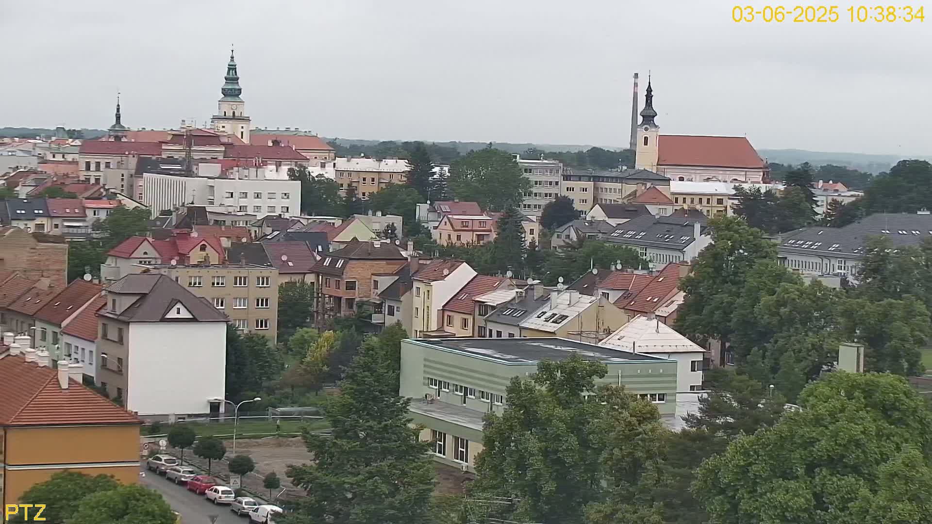 A European town under an overcast sky, featuring a mix of residential buildings, church steeples, and lush green trees.