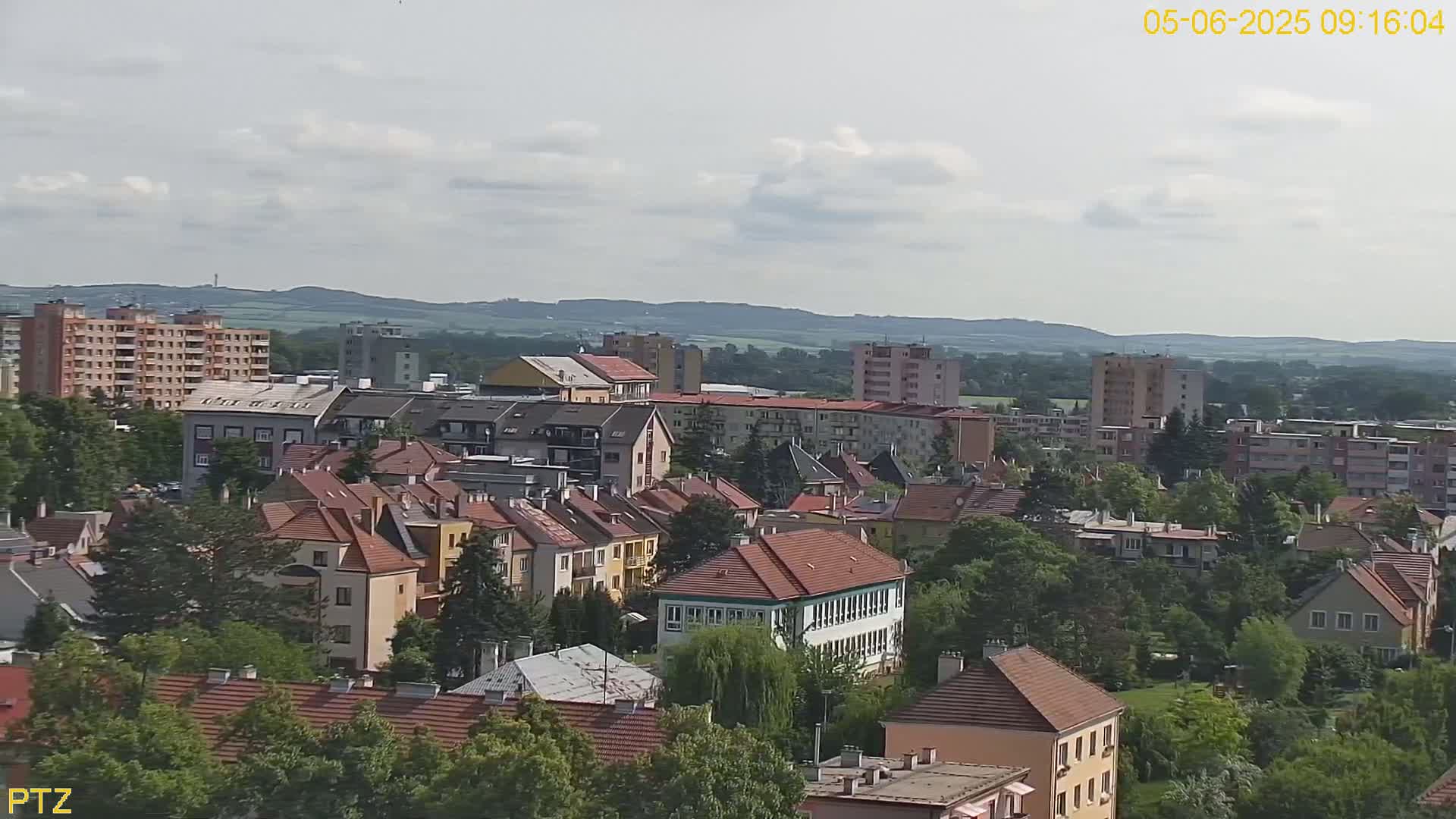 A partly cloudy day reveals a sprawling residential area of multi-story buildings and houses, nestled amidst green trees, with rolling hills visible in the distance.