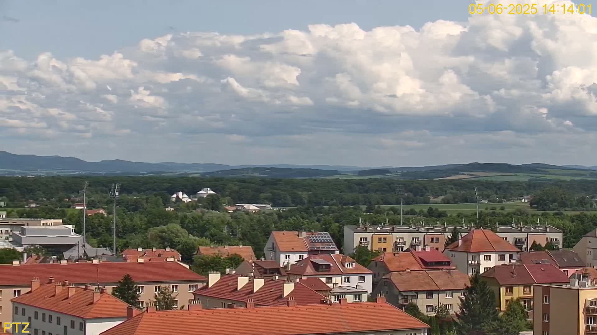 A partly cloudy sky overlooks a town of red-roofed houses nestled in a green valley, with distant rolling hills in the background.