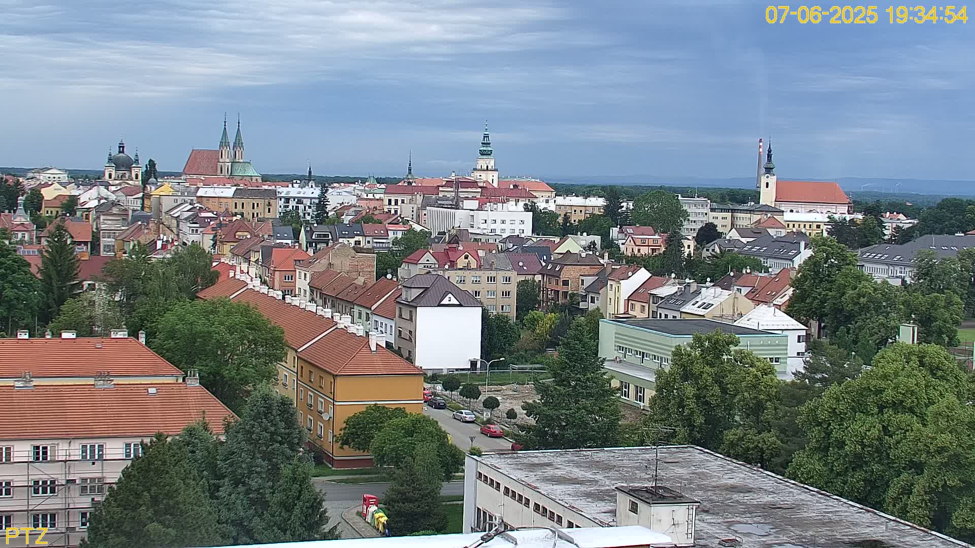 A sprawling town with numerous red-roofed buildings, interspersed with green trees, under a partly cloudy sky.