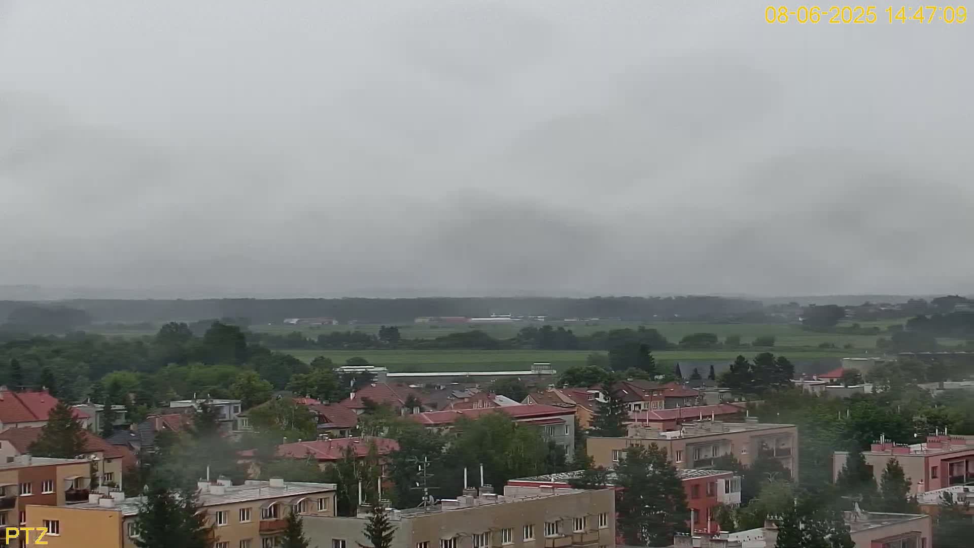 A hazy, overcast sky hangs over a residential area, which sits beside a flat expanse of green fields and trees.
