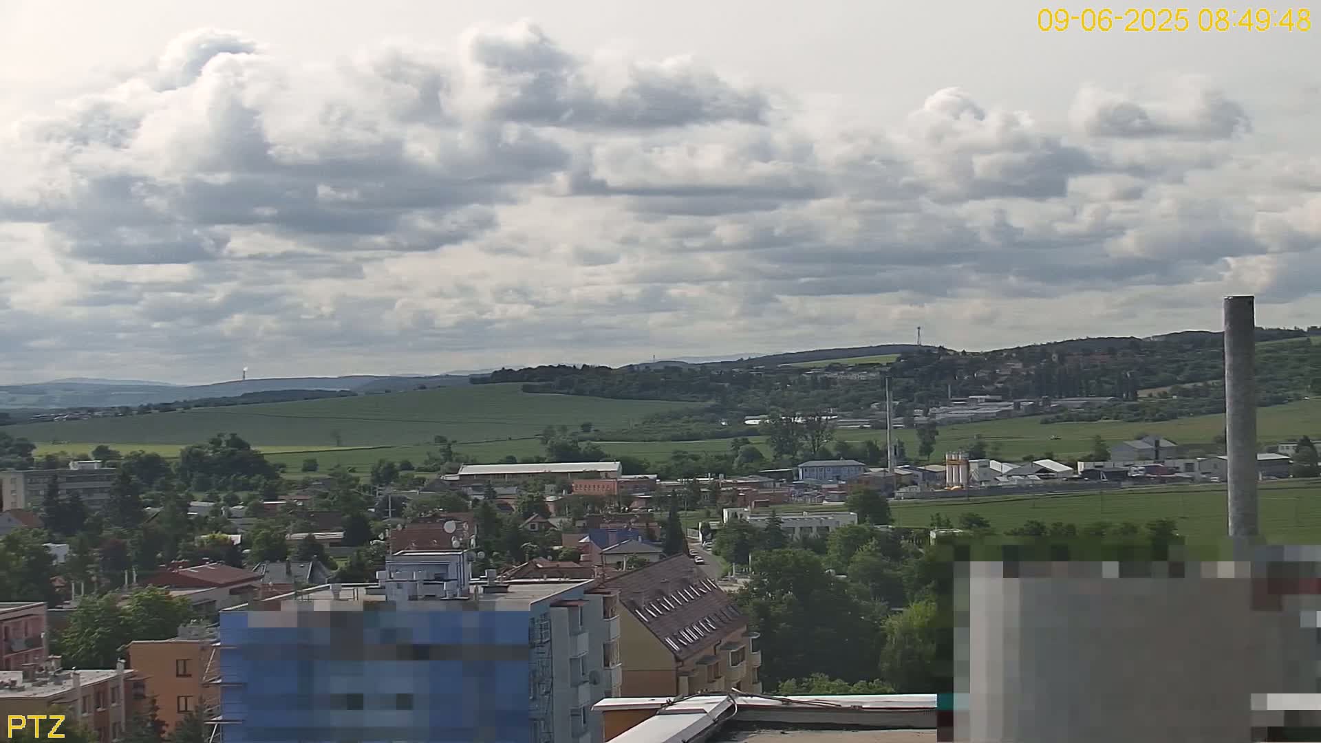 A partly cloudy day reveals a view of a town nestled amongst rolling green hills, with a tall industrial chimney visible in the distance.