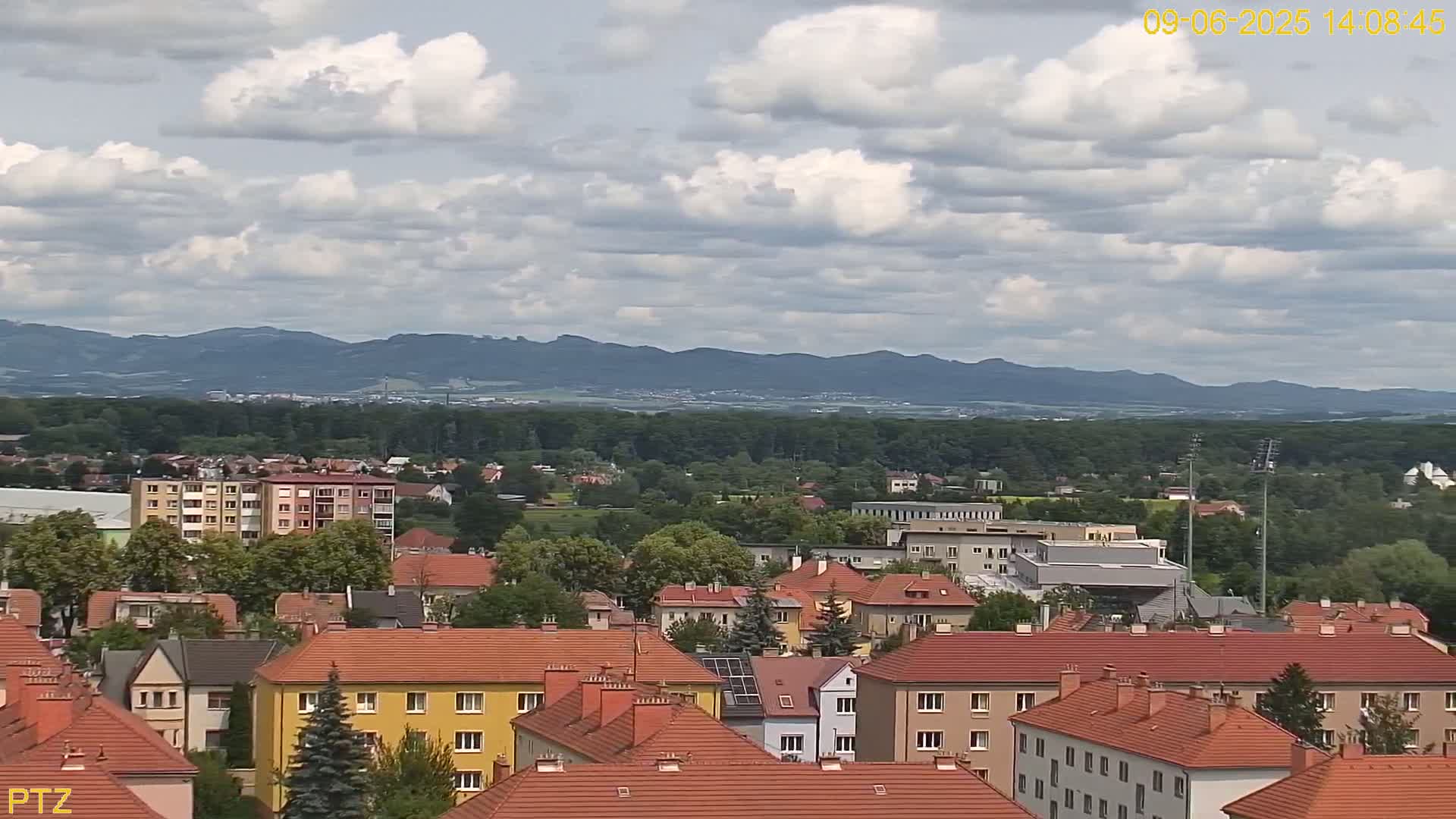 A partly cloudy day reveals a view of a town nestled among green trees and distant mountains.