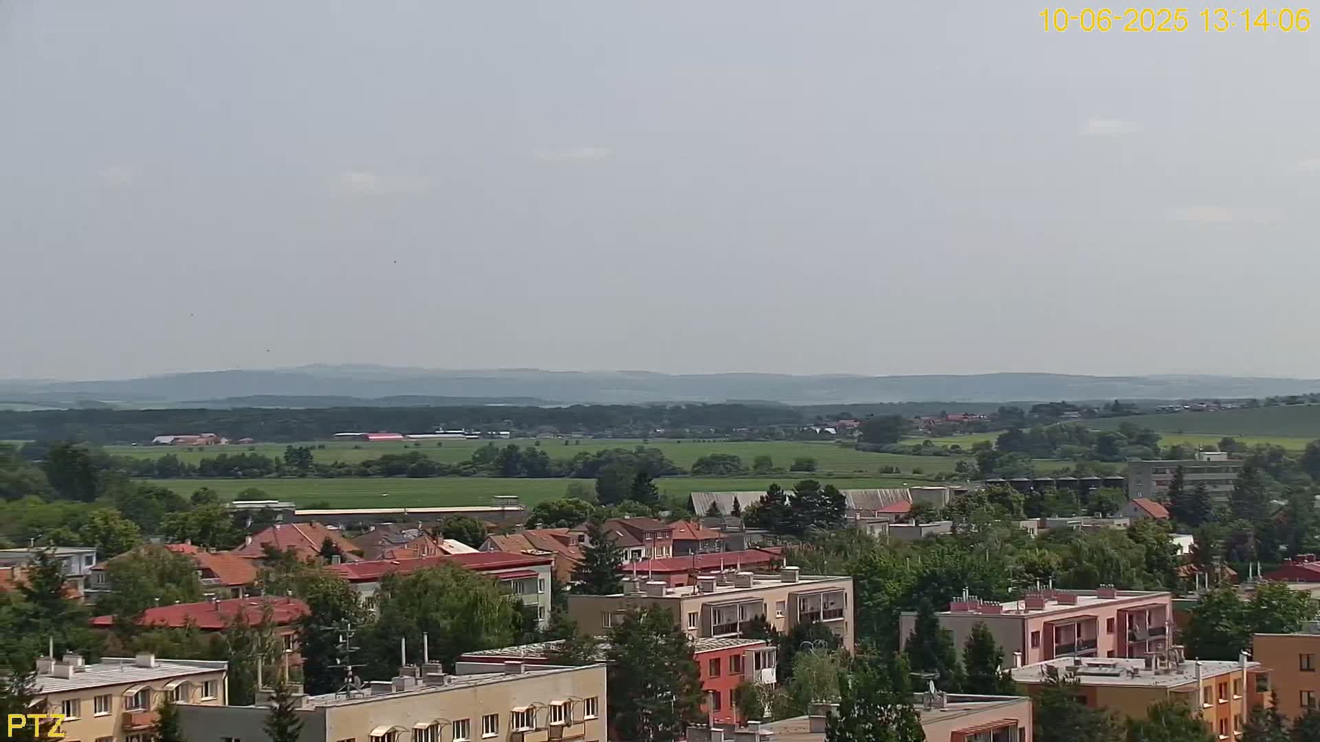 A hazy, sunny day reveals a sprawling cityscape of low-rise buildings and trees, extending to a distant, rolling green landscape under a pale sky.