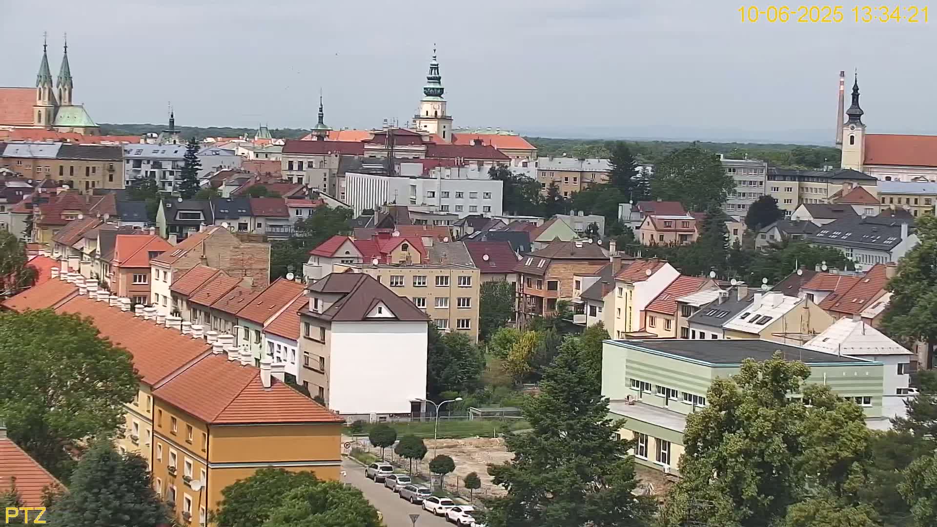 A high-angle, long shot on a sunny day shows a dense collection of buildings with red-tiled roofs, some taller buildings, churches, and a few cars on a street below.