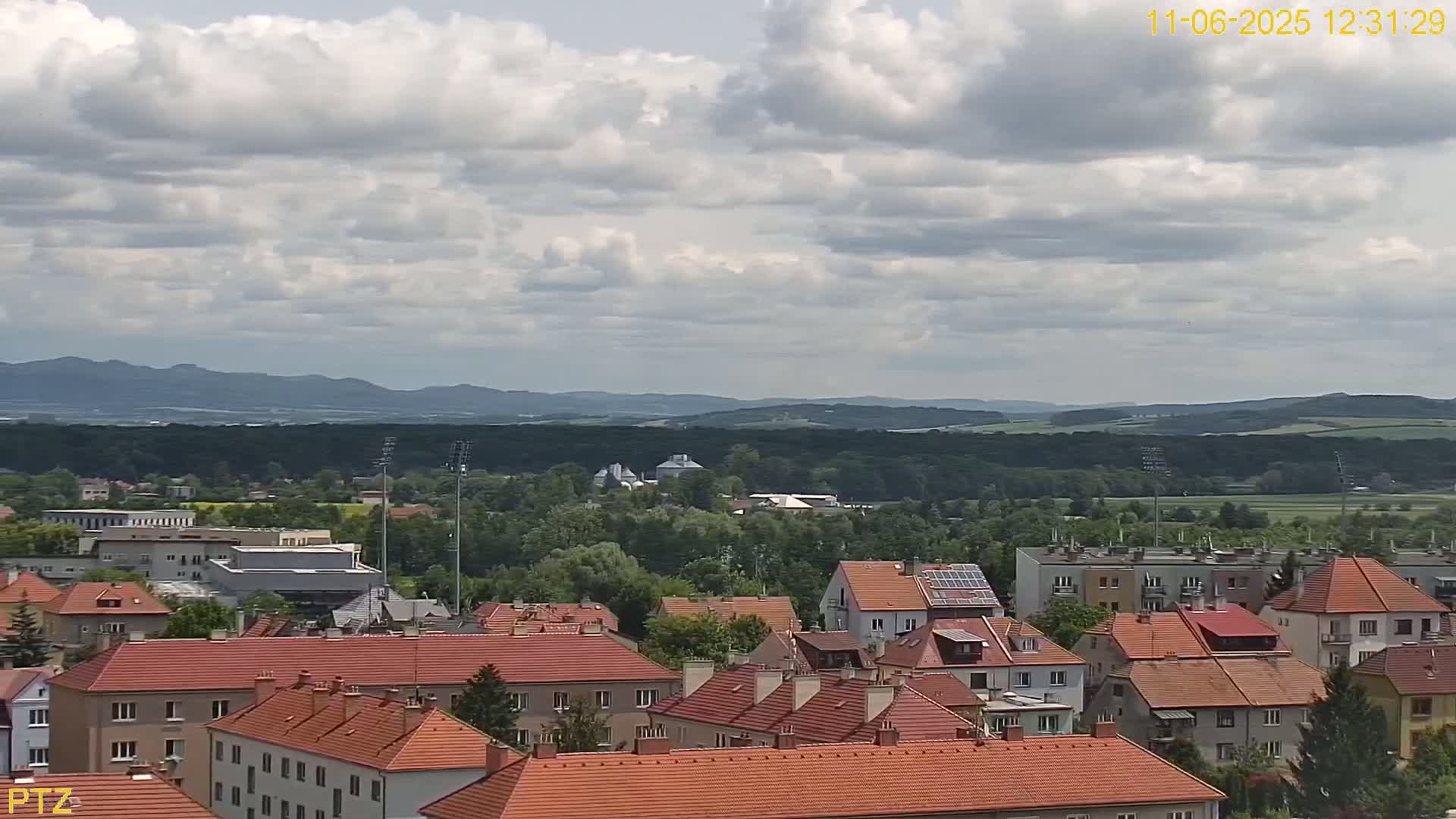 A partly cloudy day reveals a view of a town with red-tiled roofs nestled amongst green trees, backed by rolling hills and distant mountains.