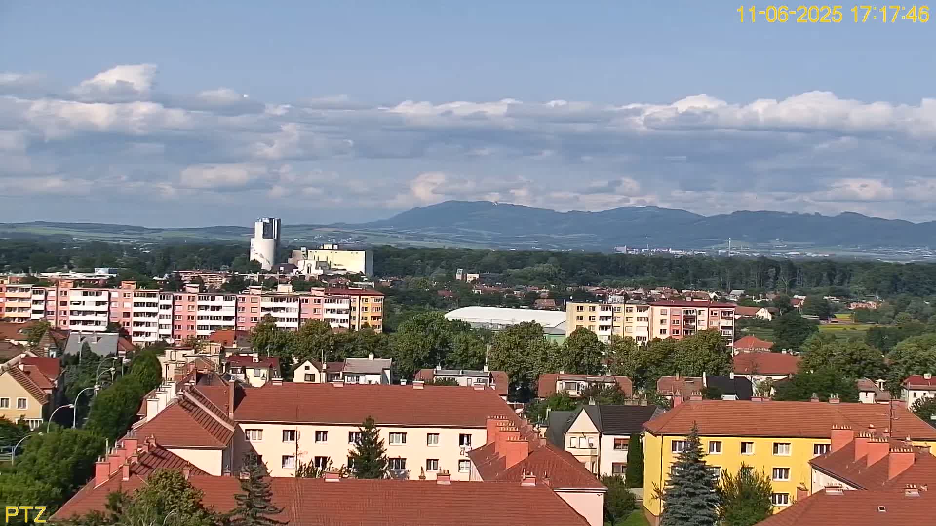 A partly cloudy day reveals a cityscape of low-rise buildings and apartment complexes nestled in a valley, with mountains visible in the distance.