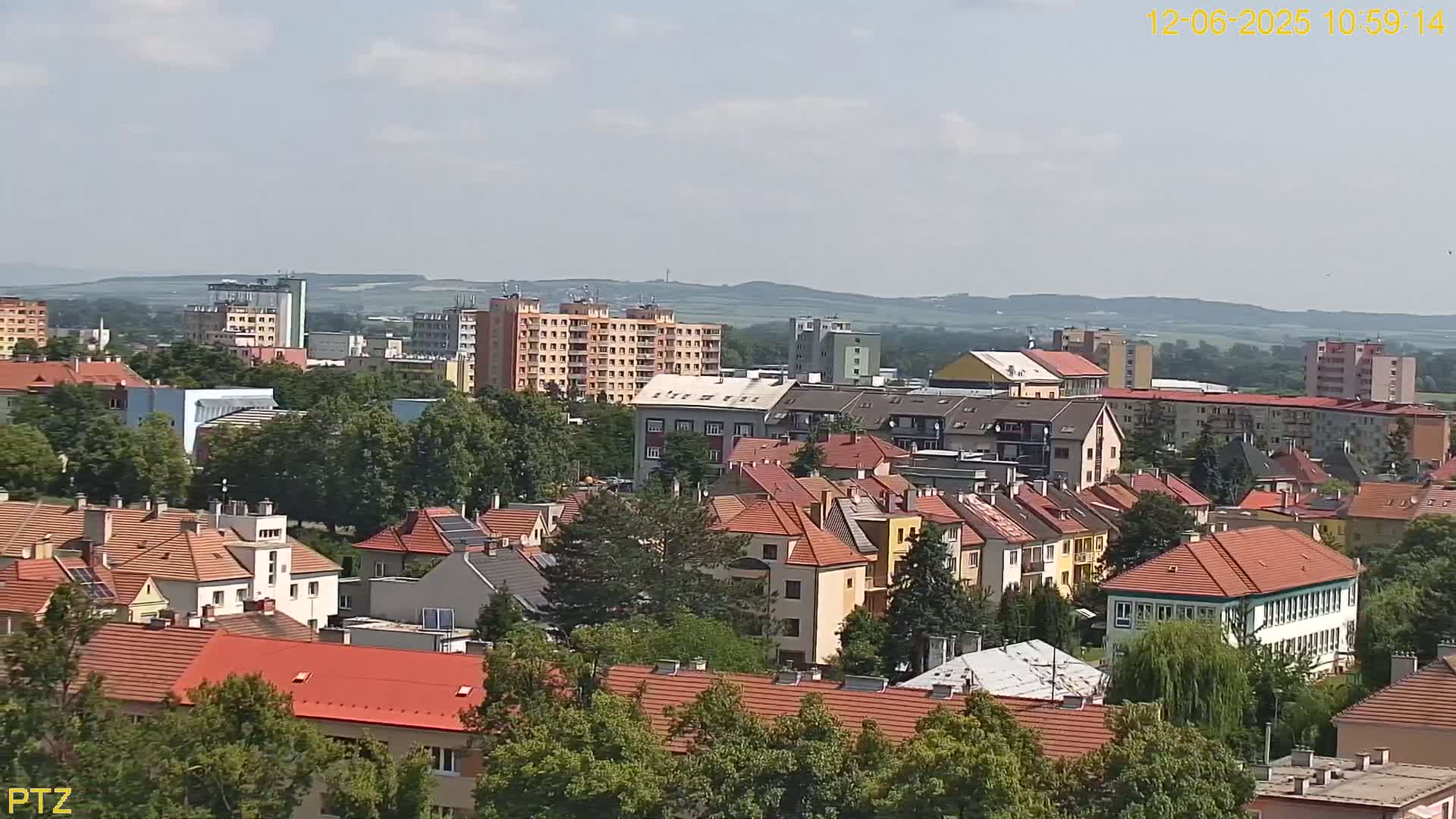 A sprawling town of multi-story buildings with red tile roofs, nestled among green trees under a partly cloudy sky.