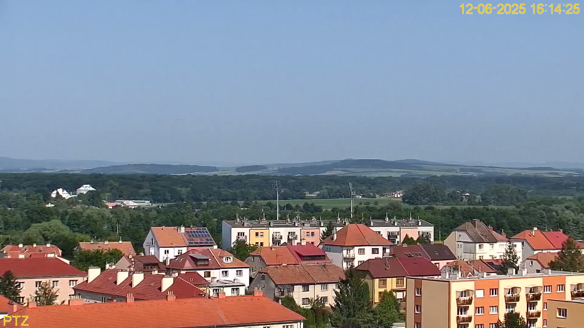 A clear, sunny day reveals a town of red-roofed houses nestled amongst green hills and forests under a pale blue sky.