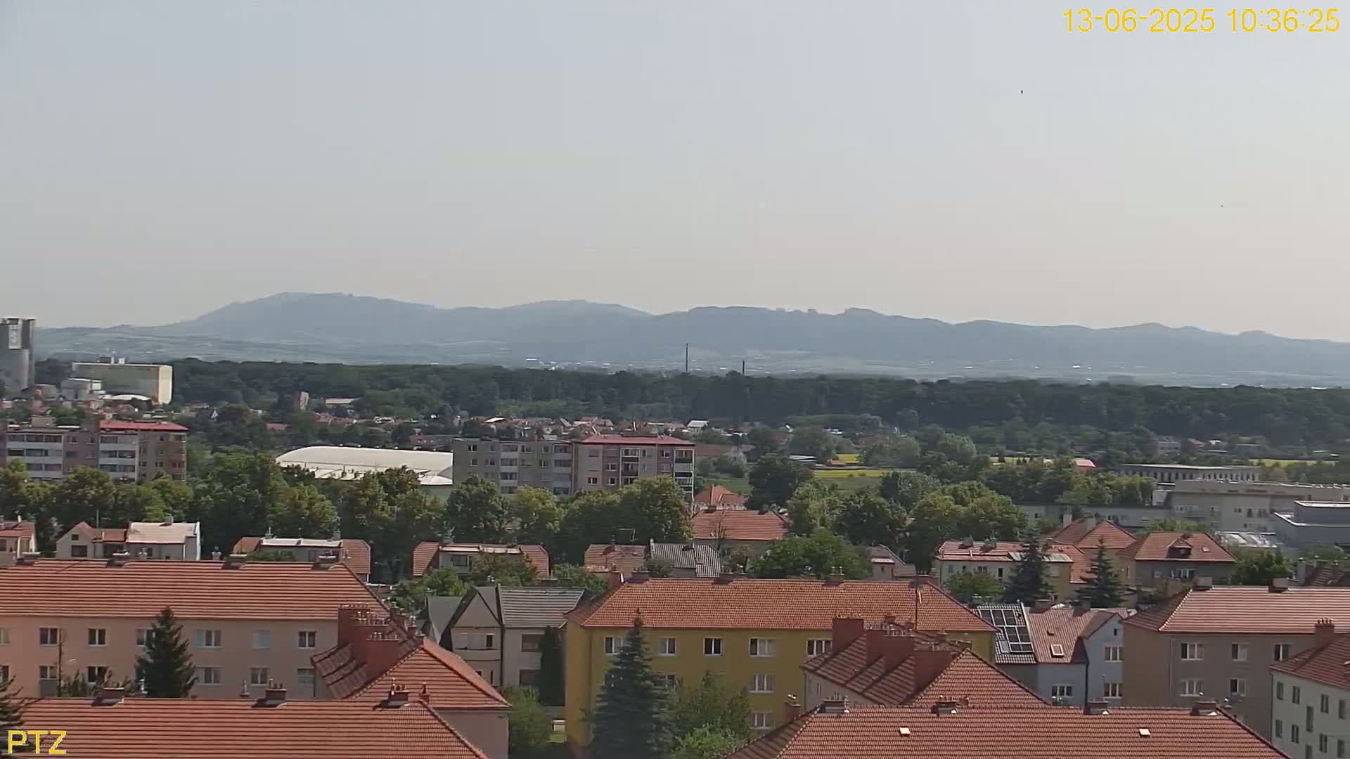 A hazy, sunny day reveals a cityscape of red-tiled roofs and multi-story buildings, with a backdrop of distant, low-lying mountains and a vast expanse of green forest.