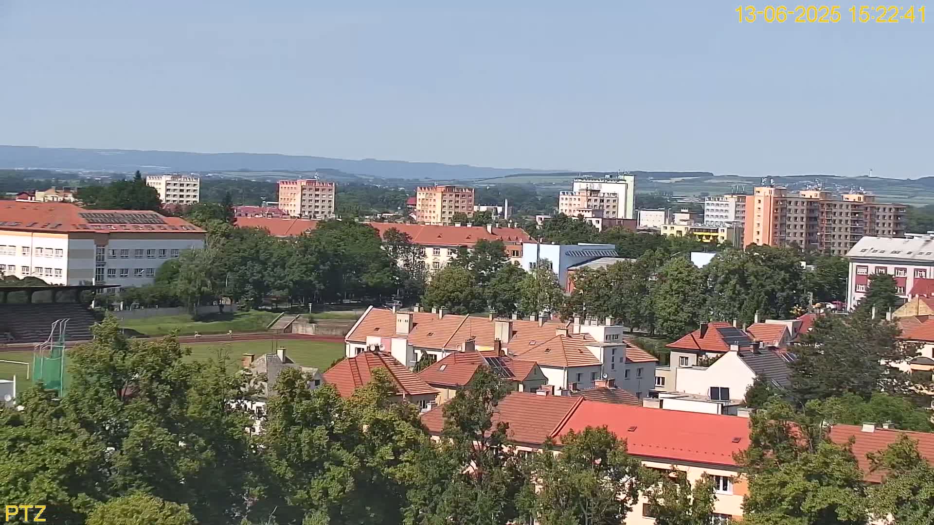 An aerial view of a town on a sunny day shows numerous residential buildings, a school, and a sports field surrounded by lush green trees.