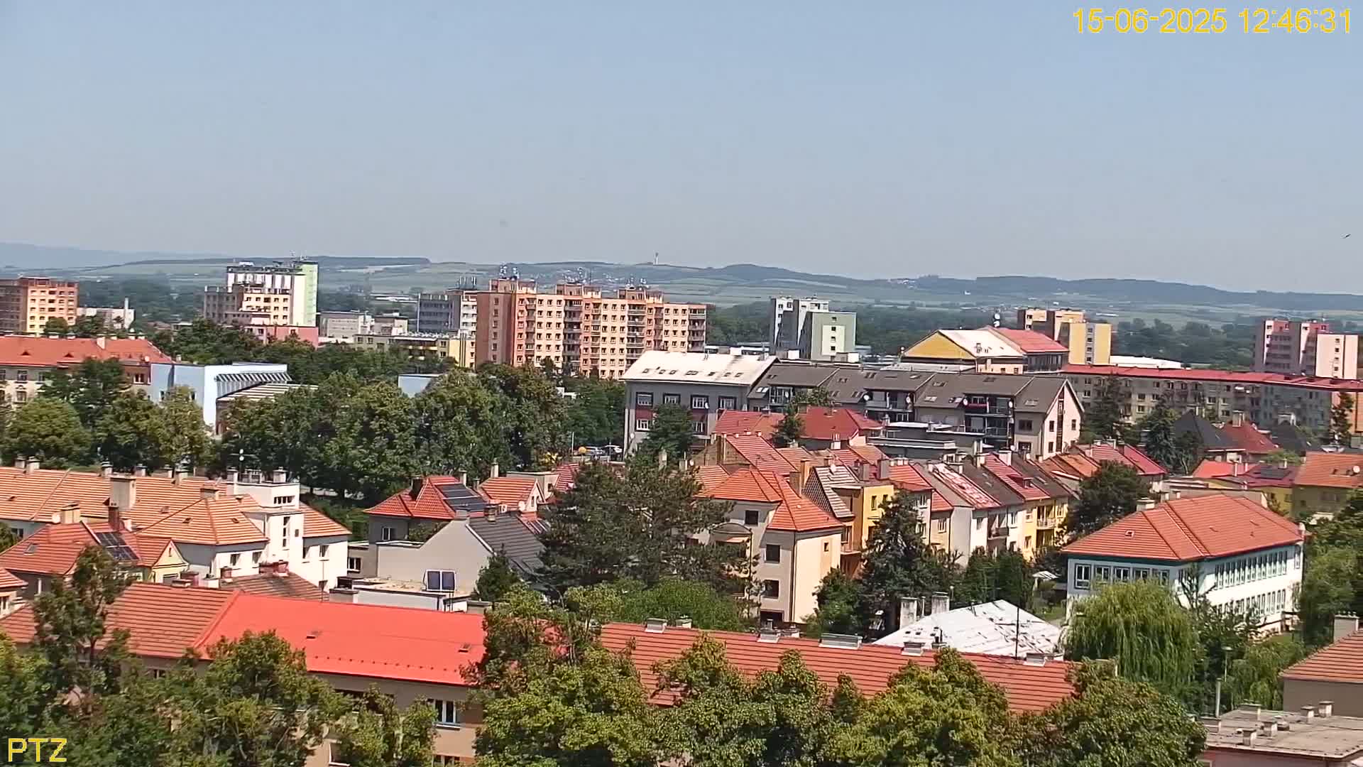 A high-angle, long shot on a clear, sunny day shows a cityscape of many low-rise buildings with red-tiled roofs and interspersed taller buildings, all surrounded by green trees.