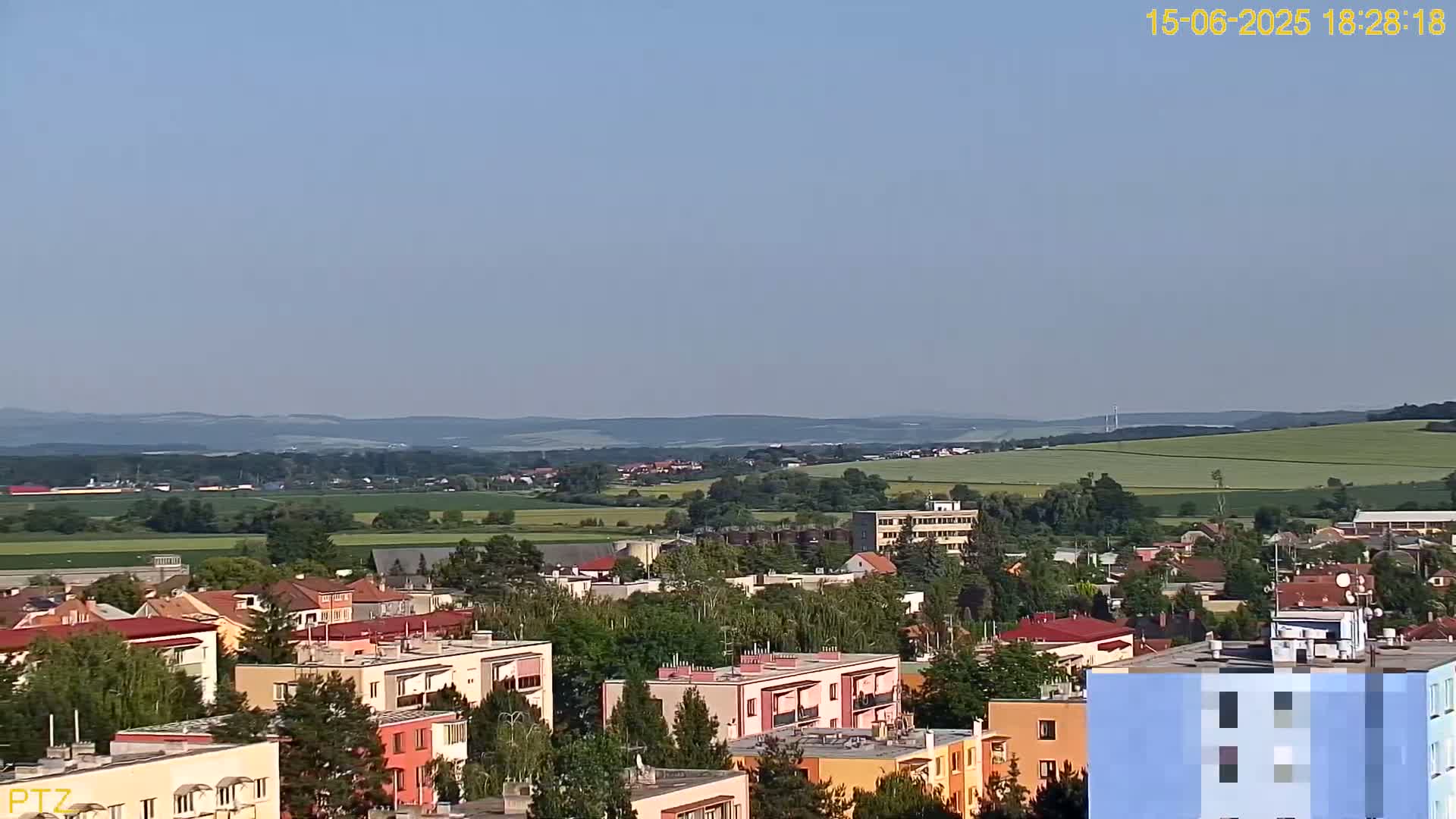 A sprawling town of low-rise buildings and apartment complexes extends into a gently rolling green landscape under a clear, light-blue sky.
