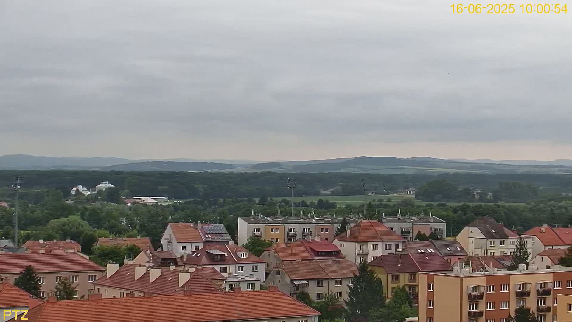 A view of a town with red-tiled roofs nestled amongst green trees, under a cloudy sky, with distant rolling hills in the background.
