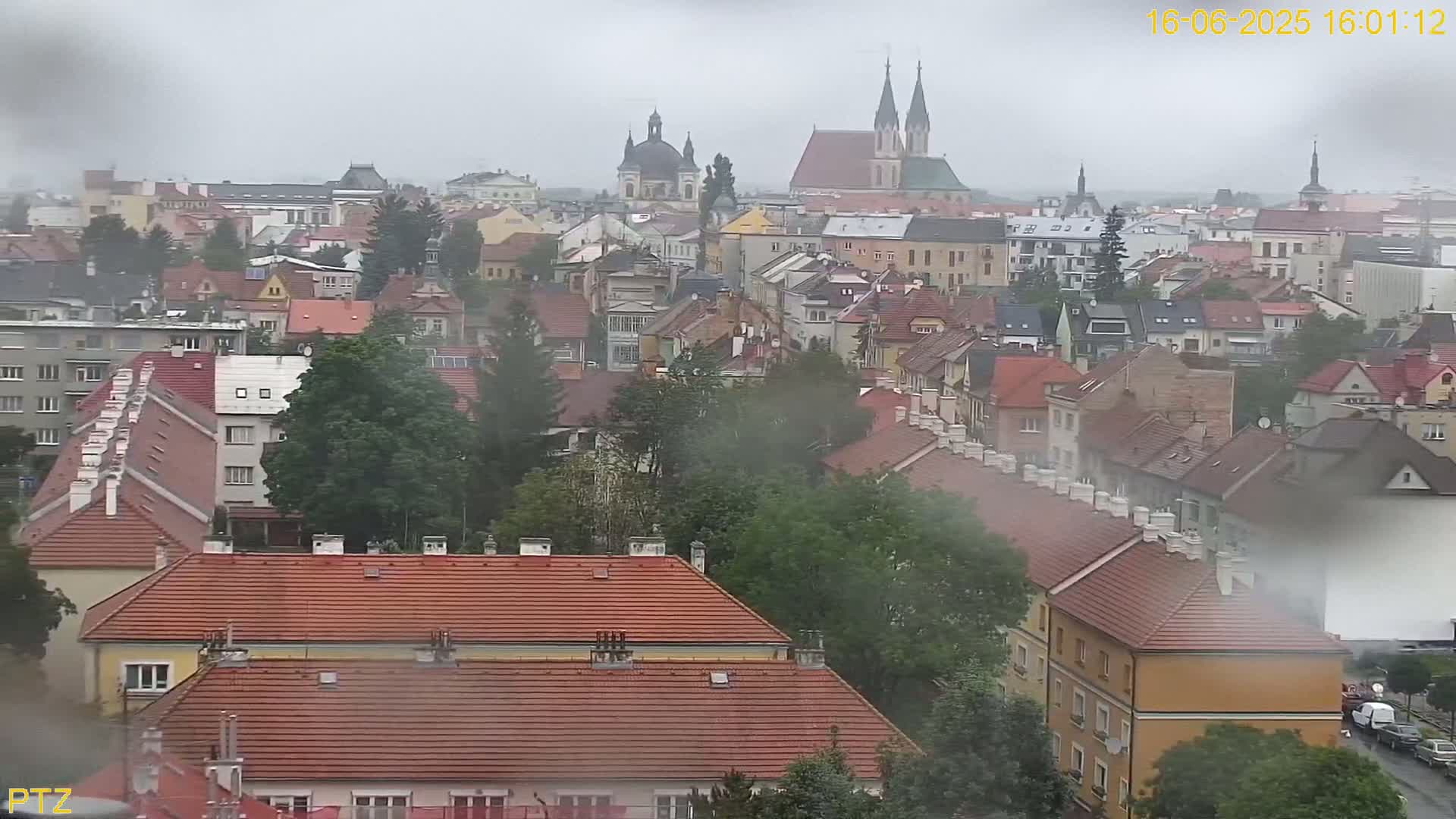 A hazy, rainy view of a European city, featuring numerous red-roofed buildings and prominent church spires in the distance.