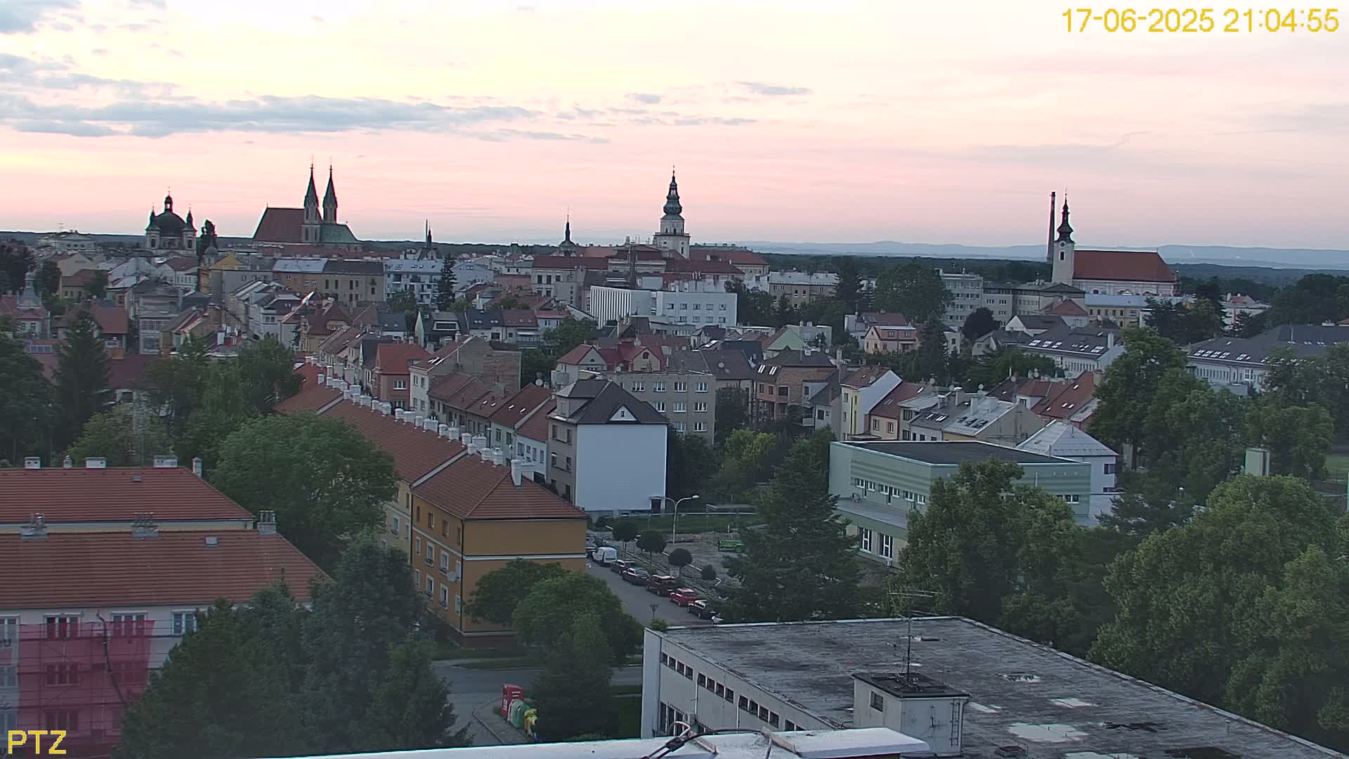 An aerial view of a European town at dusk, showing numerous buildings, churches, and trees under a pinkish sky.