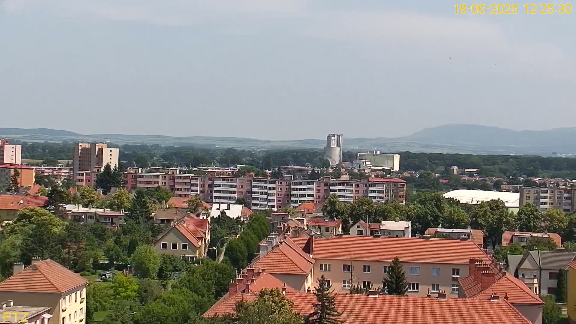 A residential area with many low-rise buildings and a few taller apartment buildings is shown, set against a backdrop of hills and under a mostly clear sky.