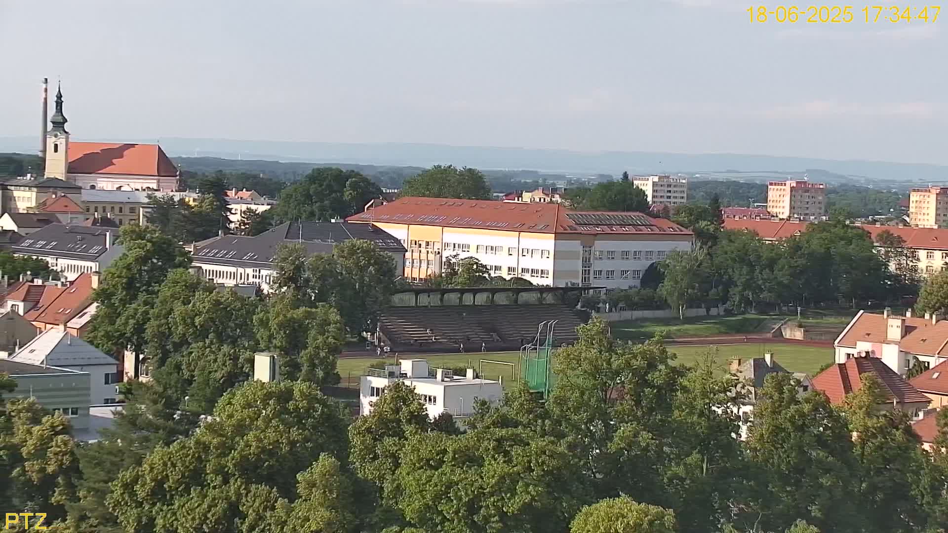An aerial view of a town on a clear, sunny day shows a large school building, a running track, and numerous buildings surrounded by lush green trees.