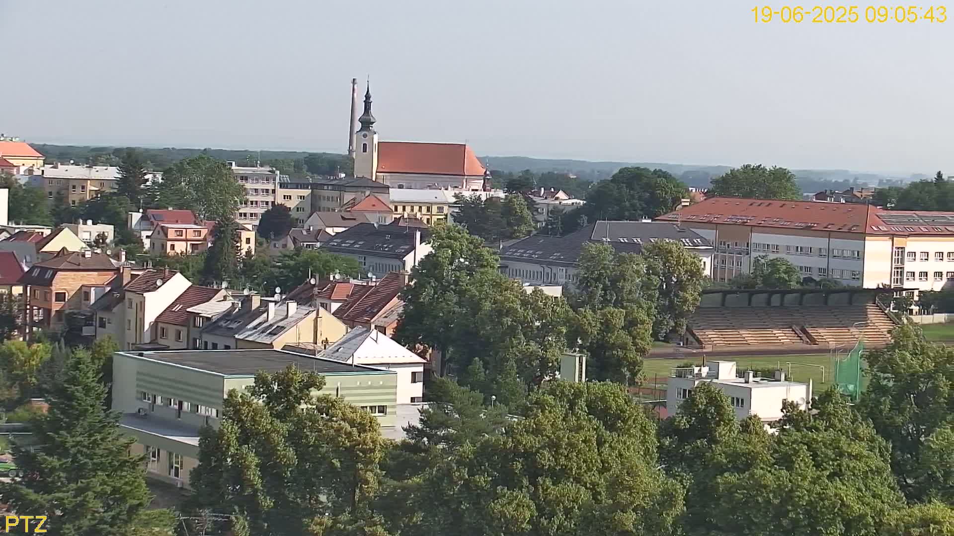 An aerial view of a town on a clear, sunny day shows many buildings surrounded by lush green trees, including a church with a tall steeple and a stadium with empty bleachers.