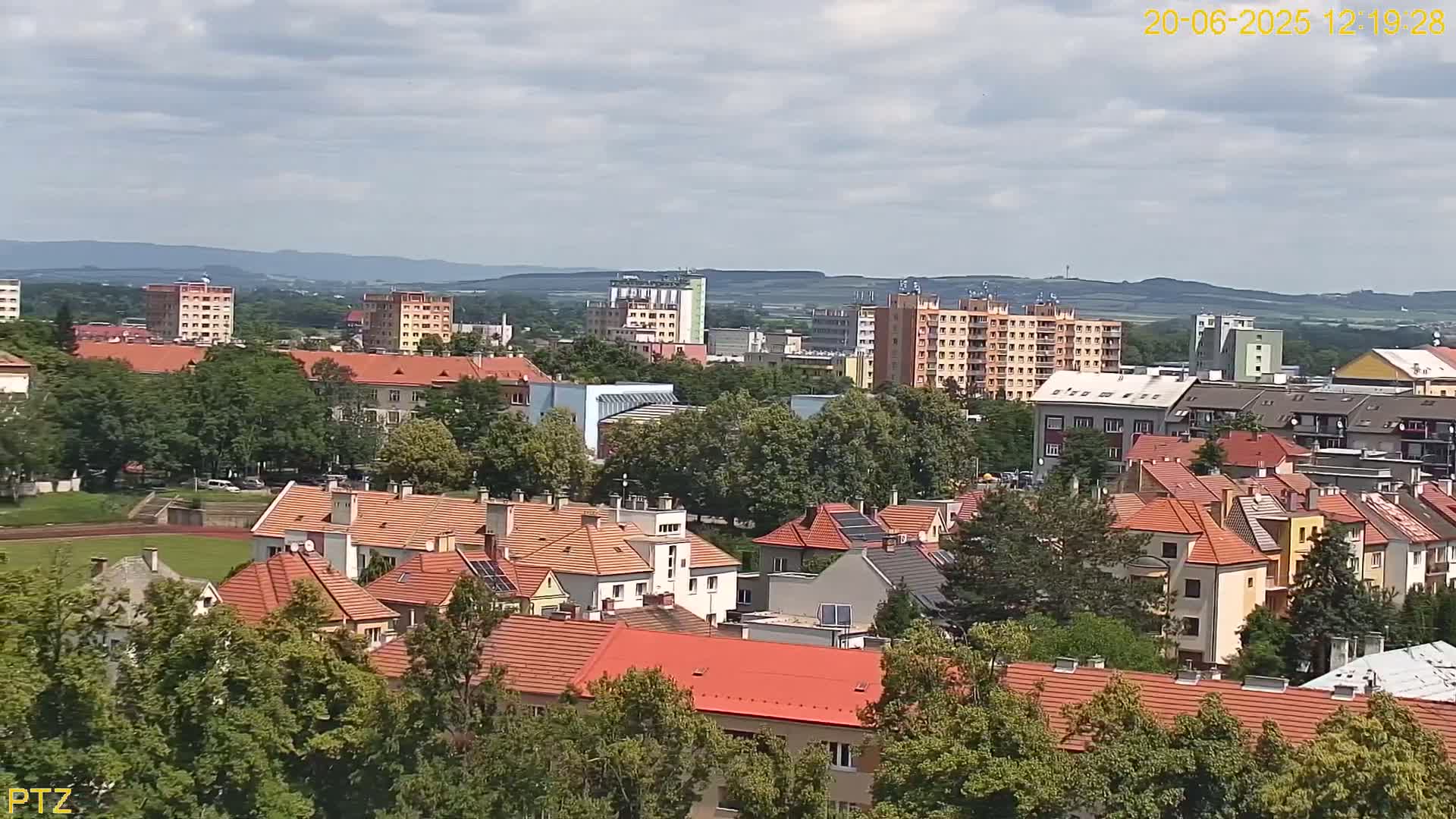 A partly cloudy day reveals a sprawling town of predominantly low-rise buildings with red tile roofs, interspersed with taller apartment buildings and surrounded by green trees and hills in the distance.
