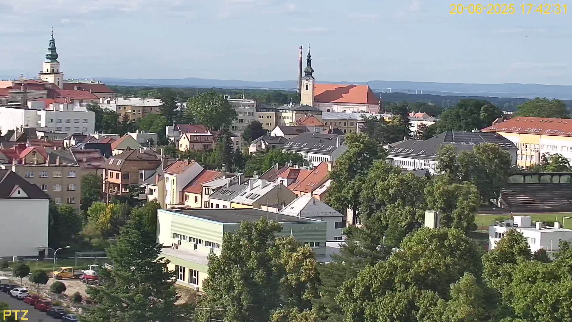 A high-angle view of a town on a partly cloudy day shows many buildings, mostly low-rise, surrounded by lush green trees, with a church steeple prominent in the distance.