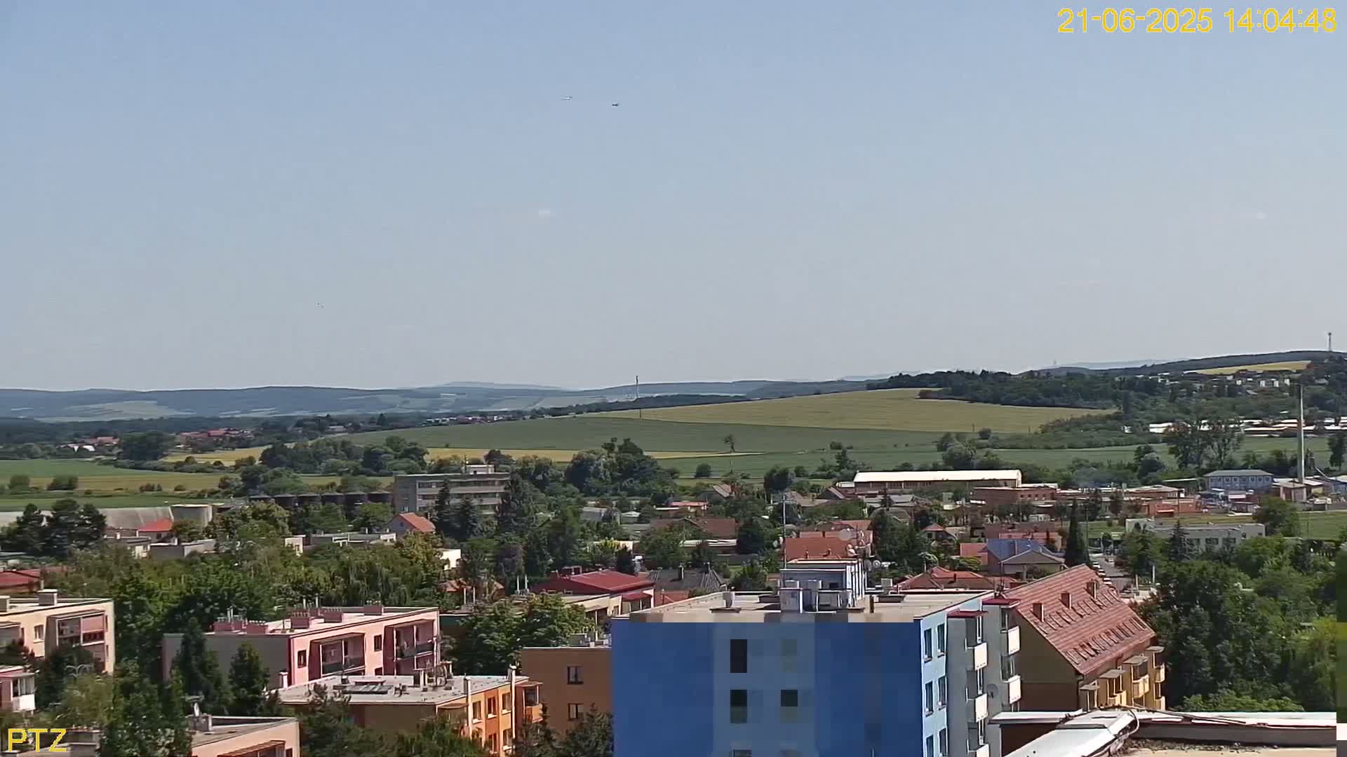 A clear, sunny day reveals a sprawling town nestled amidst rolling green hills and fields, with several multi-story buildings prominent in the foreground.