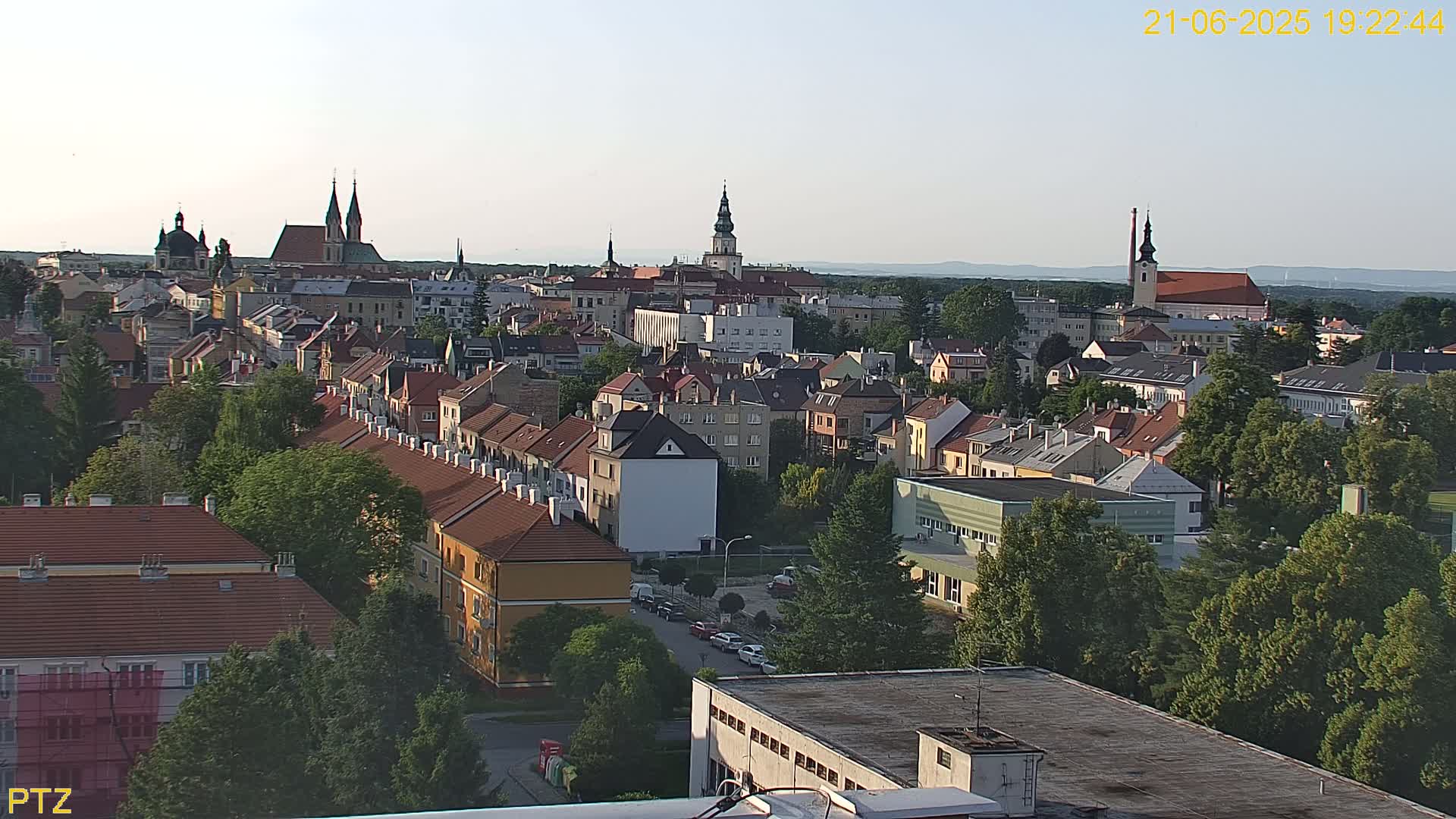 An elevated view of a European town under a clear, twilight sky, showing many red-tiled roofs, green trees, and several churches.