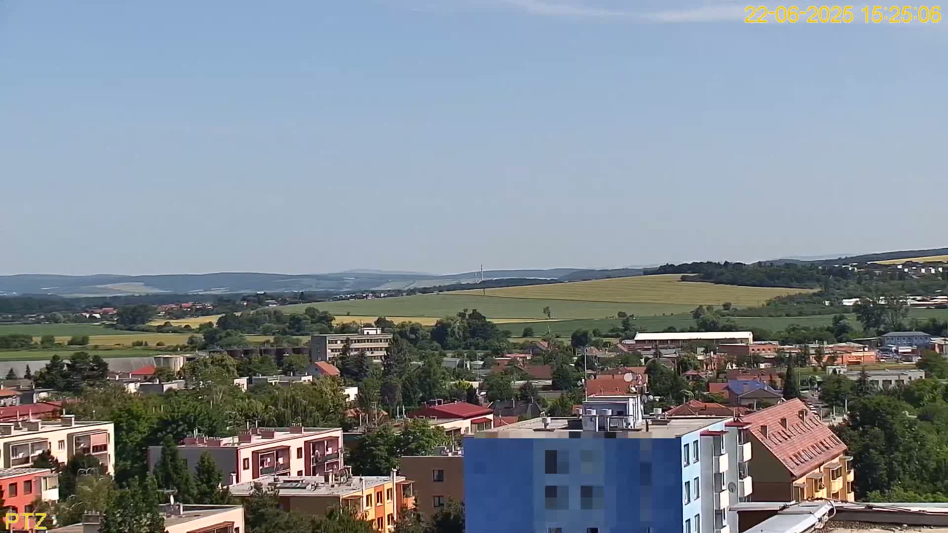 A sunny day reveals a sprawling town nestled at the base of rolling hills, with a mix of residential buildings and verdant fields stretching into the distance.