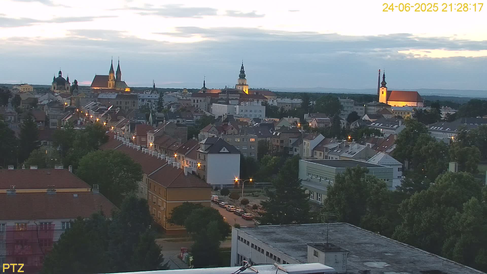 A high-angle, twilight view of a town with several church steeples and buildings illuminated, under a partly cloudy sky.