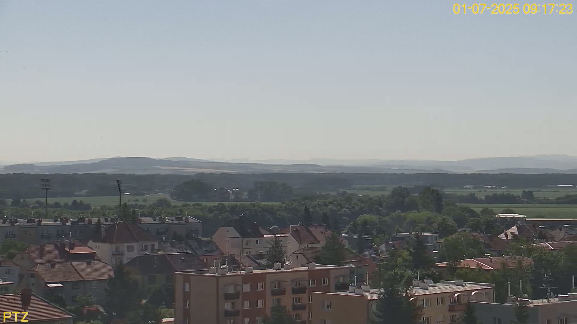 A hazy, sunny day reveals a town of low-rise buildings nestled amidst green fields and trees, with distant hills rolling in the background.