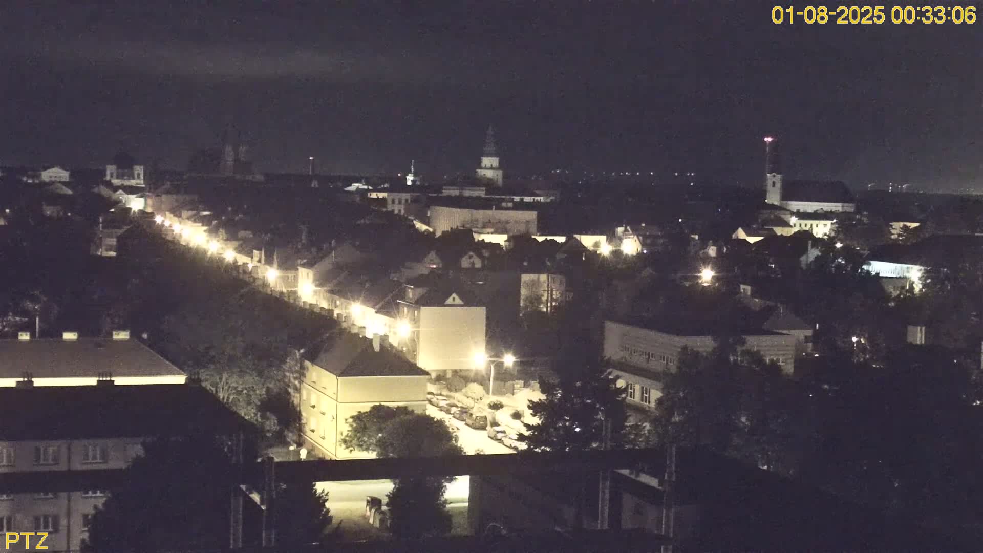 A nighttime, high-angle view of a town's streetlit buildings and roads, with a church spire visible in the distance.
