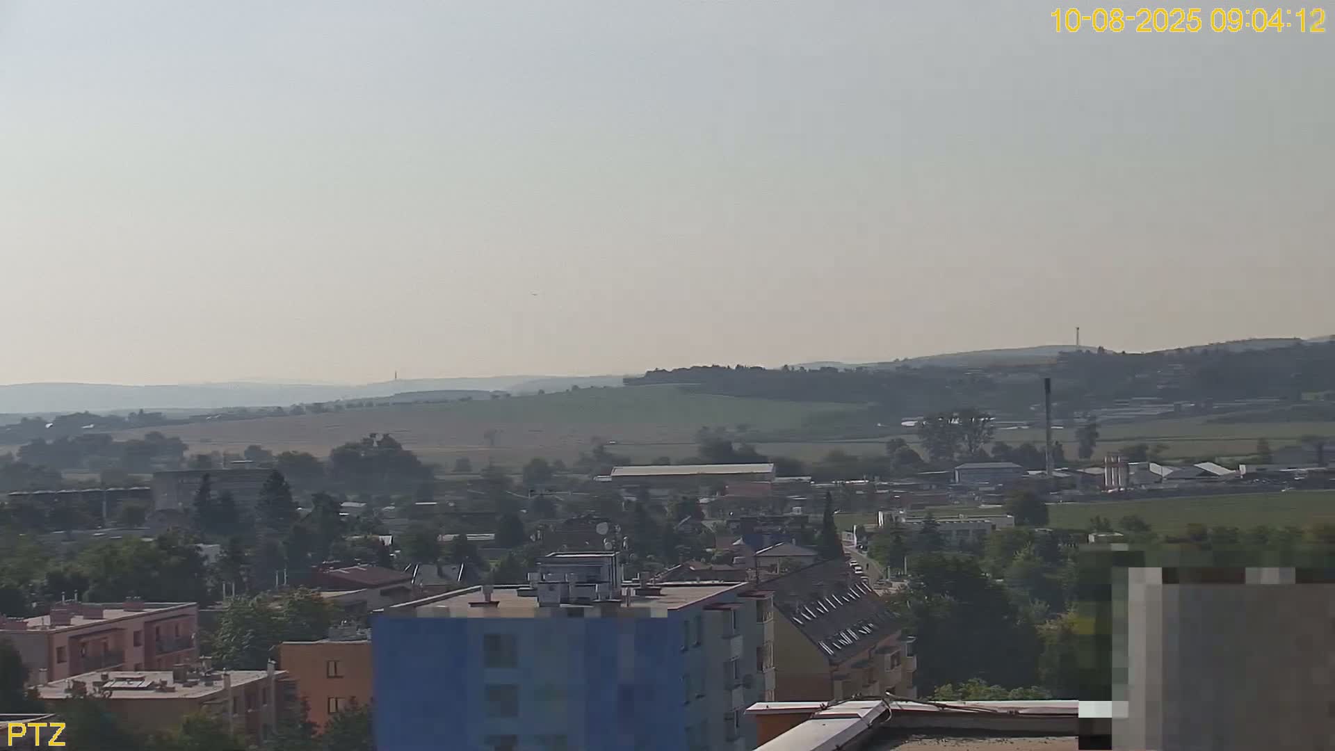A hazy, daytime view shows a town with buildings and trees, extending to rolling hills in the background.