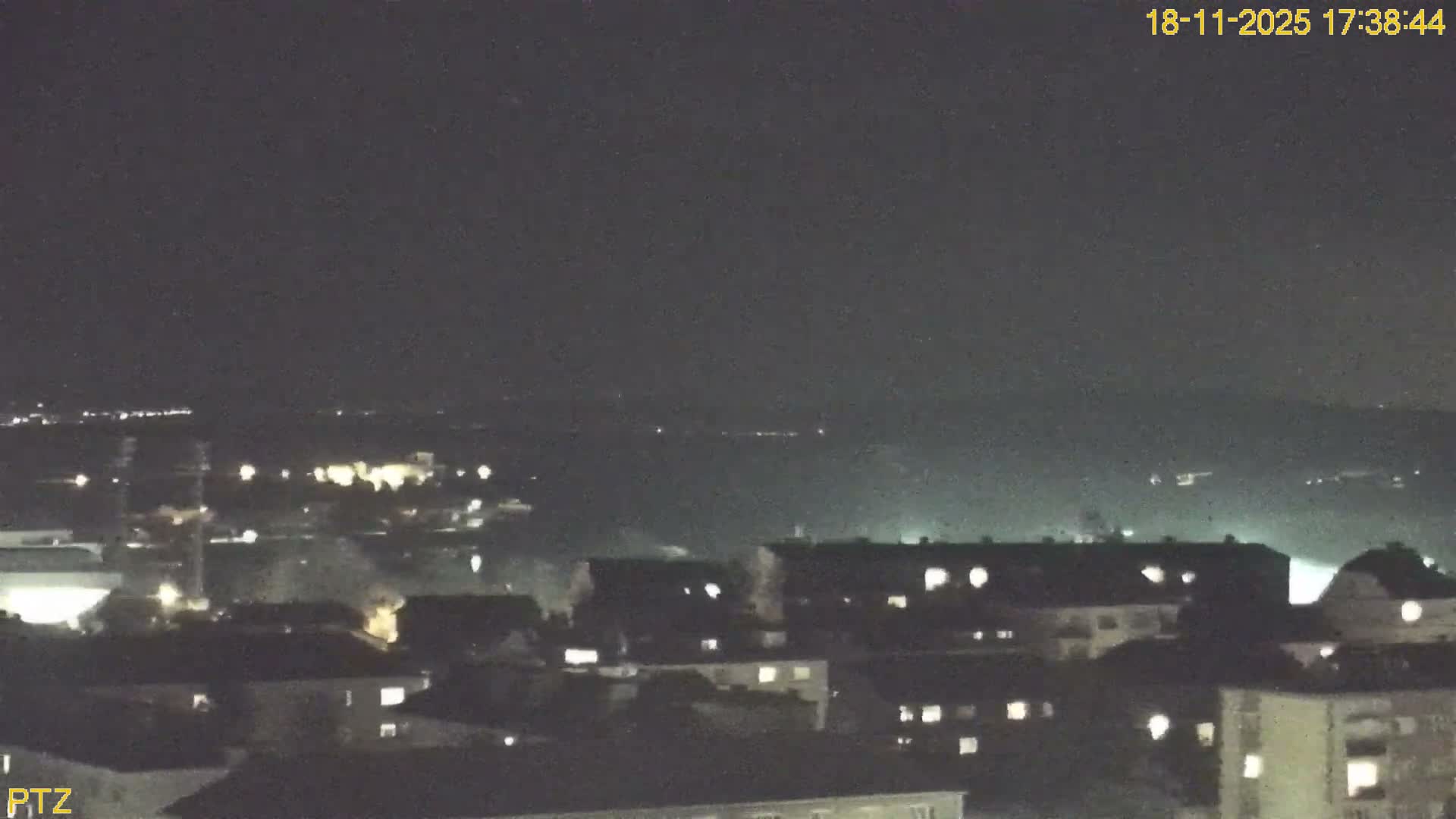 A hazy, daytime view shows a town with buildings and trees, extending to rolling hills in the background.