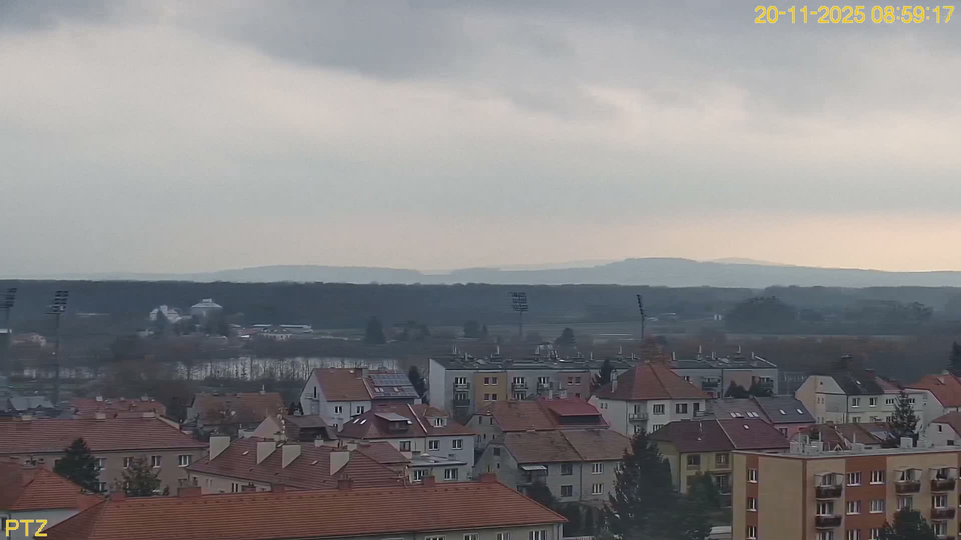 Under an overcast sky, the image displays an expansive view of a town featuring a mix of residential buildings and apartment blocks, transitioning towards a body of water, dark forest, and hazy distant hills on the horizon.