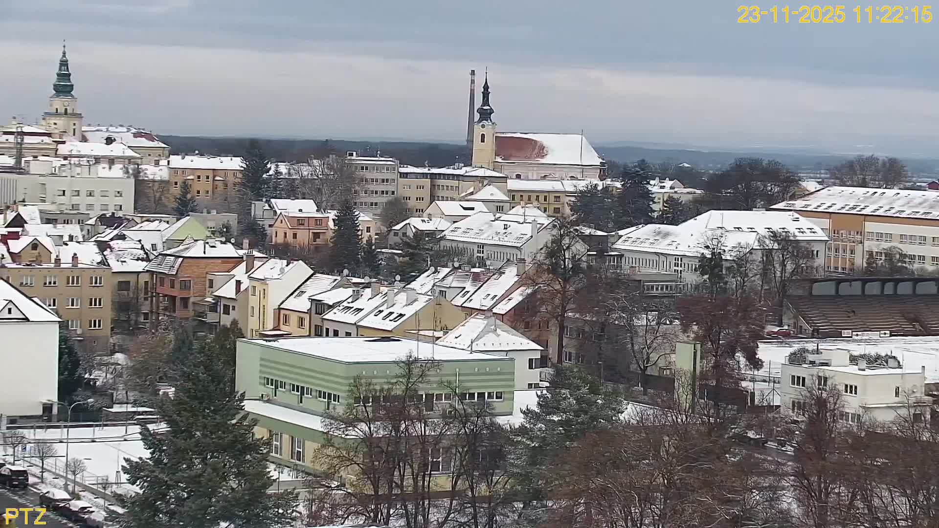 An overcast winter day reveals a snow-dusted town featuring numerous buildings with snow-capped roofs, prominent church spires, and a mix of bare and evergreen trees under a grey sky.