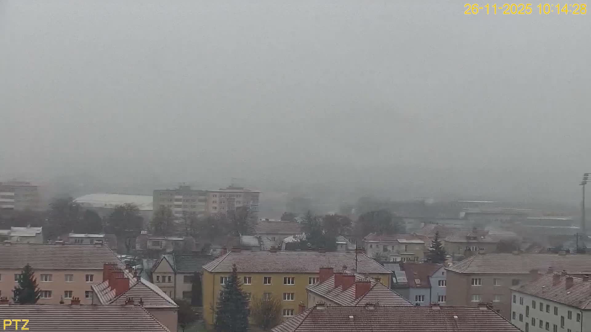 An overcast and foggy elevated view reveals a town with numerous residential buildings sporting lightly snow-dusted roofs and bare trees, with distant structures obscured by the low visibility.