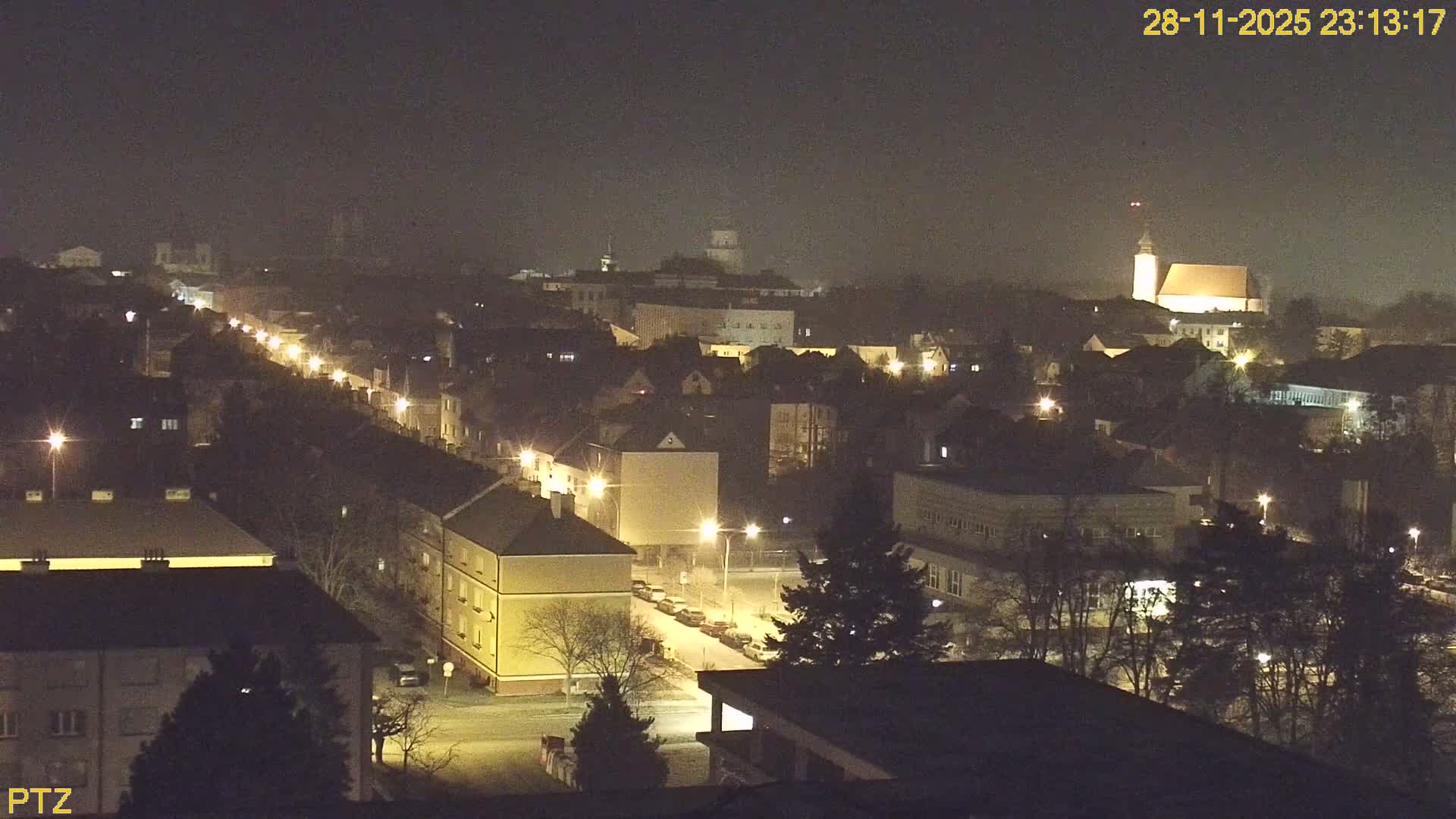 A hazy nighttime cityscape reveals numerous illuminated buildings and streets under foggy conditions, with a prominent church steeple and other distant structures faintly visible through the low visibility.