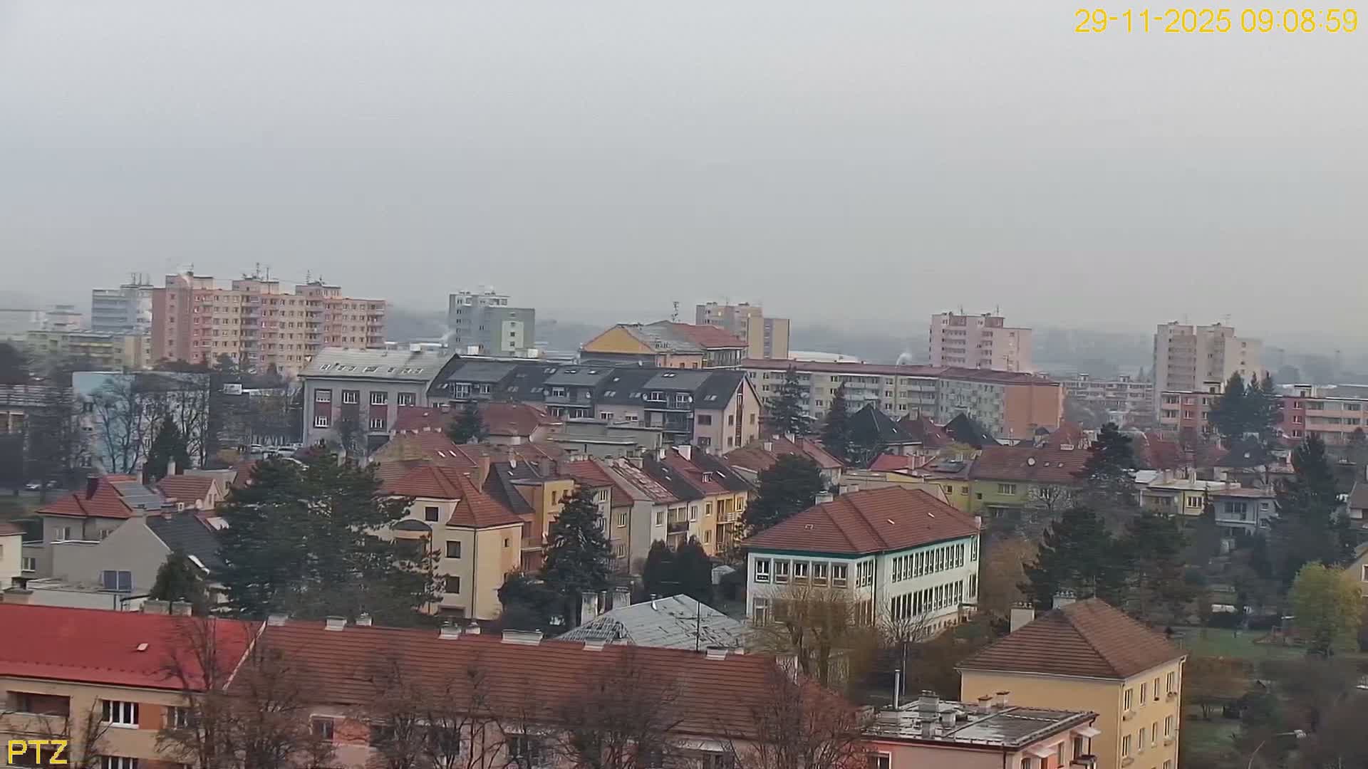The image displays an expansive view of a European town featuring a mix of multi-story apartment buildings and smaller houses with red-tiled roofs, interspersed with bare and evergreen trees, all under an overcast and hazy sky.