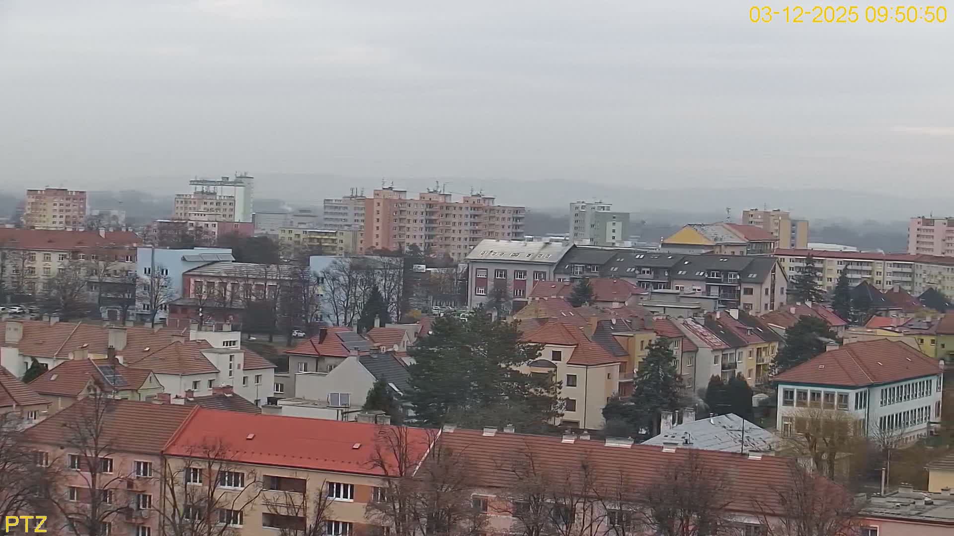 An elevated view reveals a dense urban landscape with a mix of multi-story buildings and houses with pitched roofs, all under a dull, heavily overcast, and hazy sky.