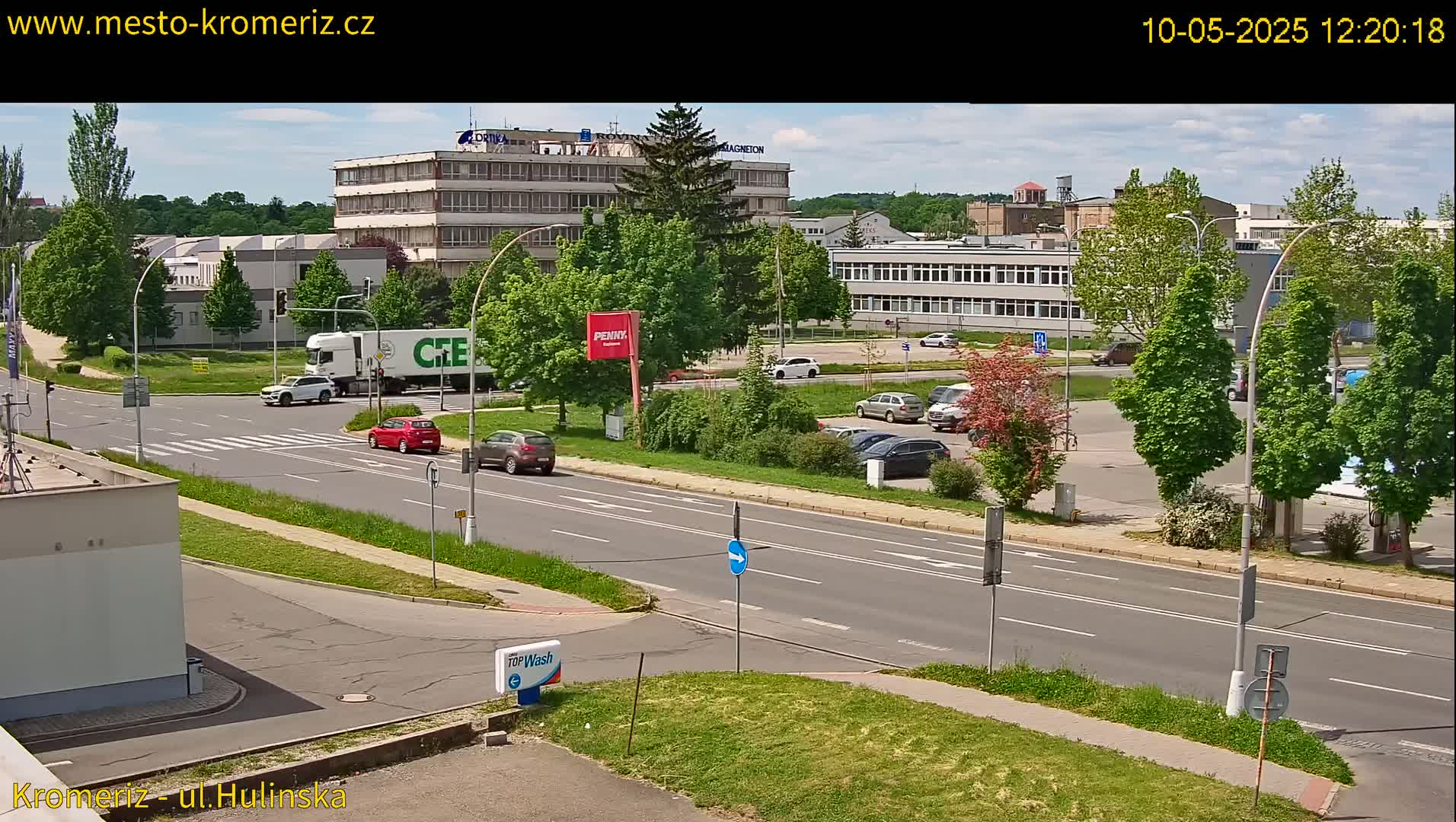 A sunny day reveals a street scene with several cars and a large truck, near a parking lot and industrial-looking buildings surrounded by green trees and lawns.