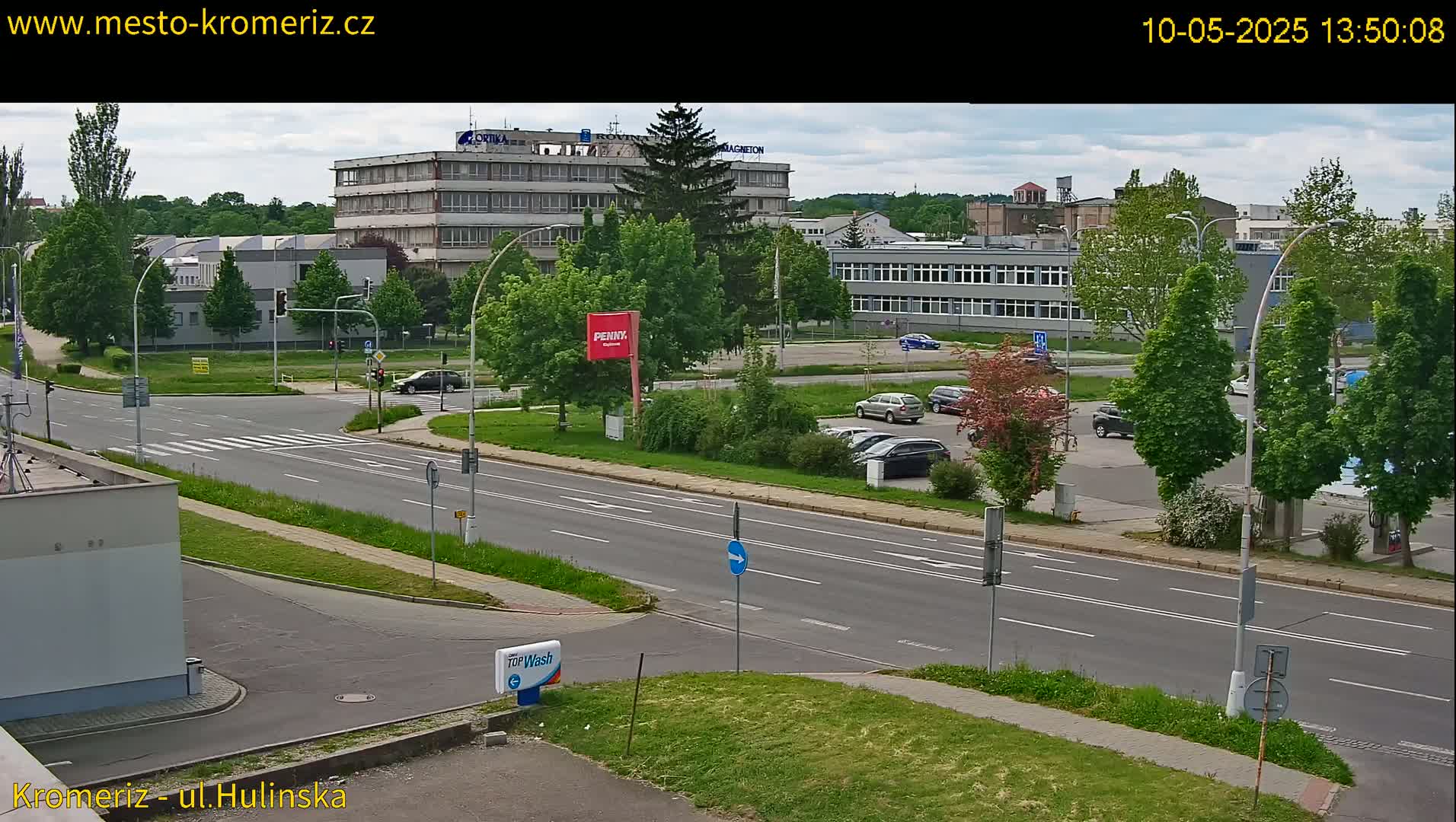 A partly cloudy day reveals a street scene with several multi-story buildings, leafy green trees, and cars parked in a lot beside a multi-lane road.