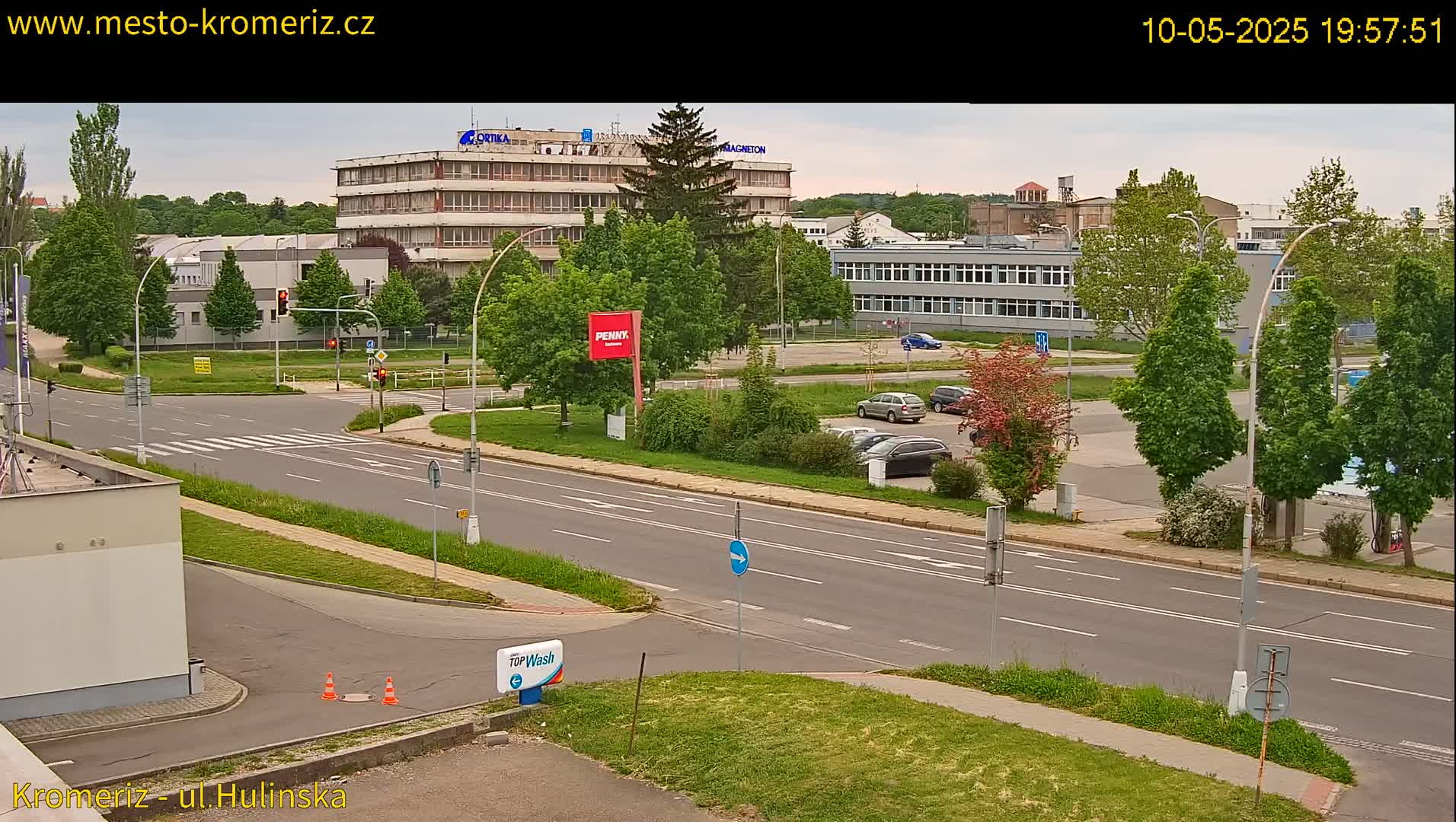 A street scene shows a multi-lane road with cars parked along the side, several buildings in the background, and green trees and grass throughout, under a partly cloudy sky.