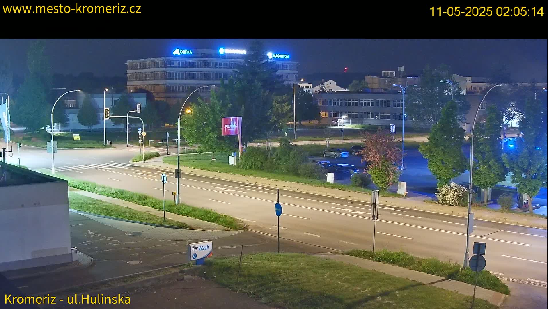 A nighttime view of a mostly empty road and parking lot, illuminated by streetlights, with several buildings in the background.