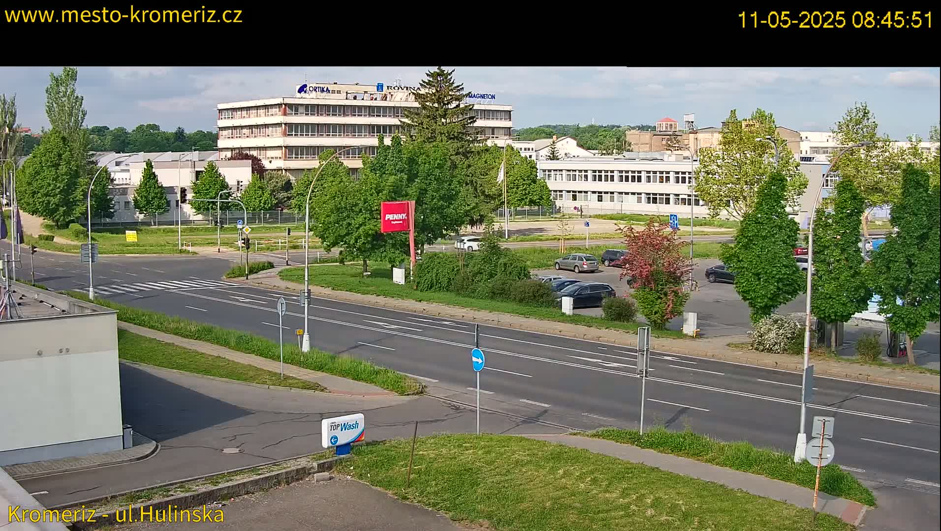 A sunny day view of a paved road intersection, with several cars parked in a lot and several buildings in the background, surrounded by lush green trees and landscaping.