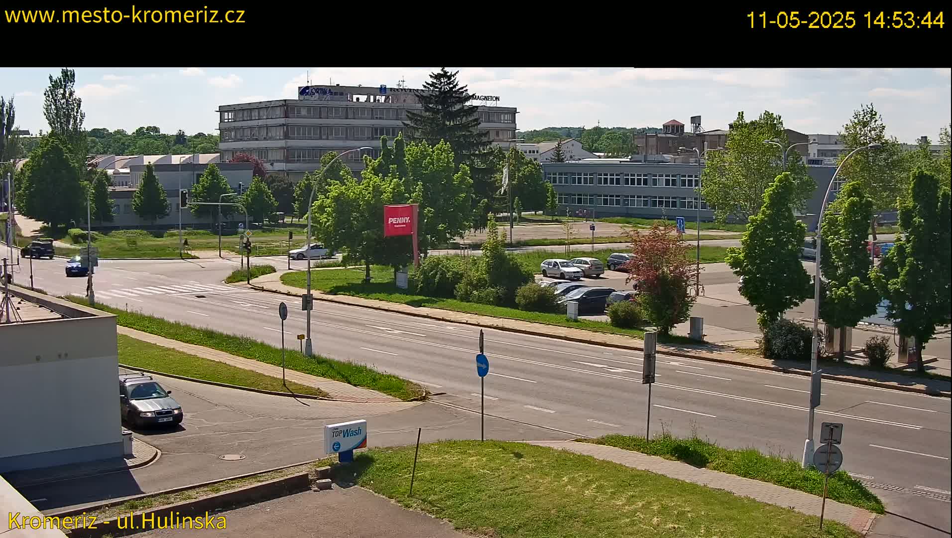 A sunny day reveals a wide road with parked cars in a lot across from several buildings, surrounded by lush green trees and grass.
