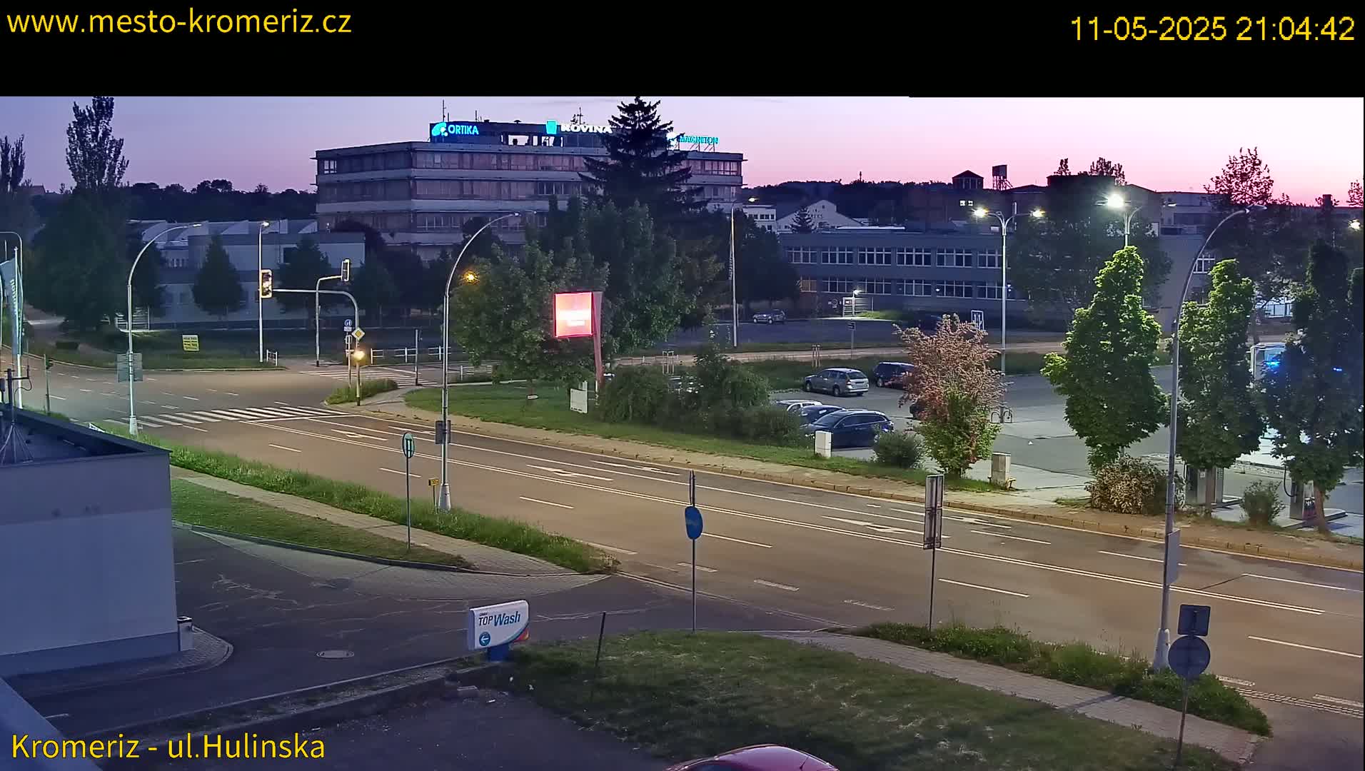 A twilight view of a multi-lane road with several cars parked in a lot and a large building in the background.